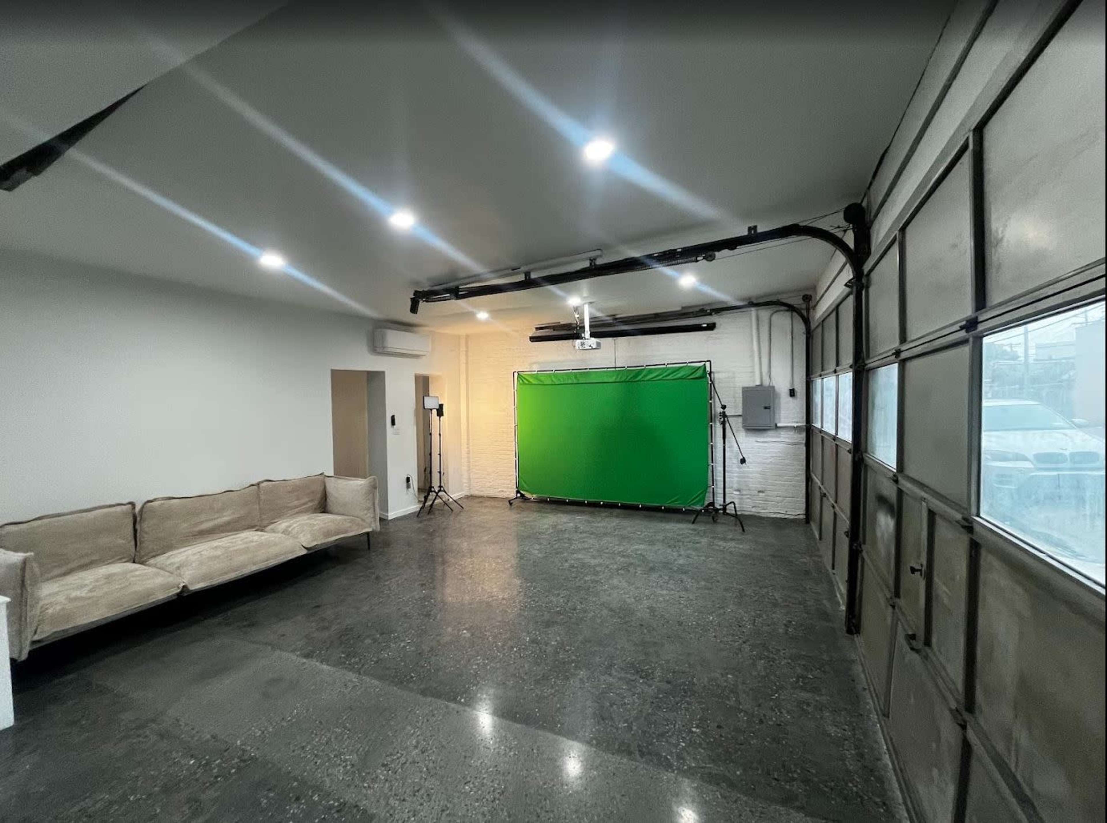 The image shows a spacious room with a green screen setup, a couch along one wall, and lighting equipment positioned nearby.