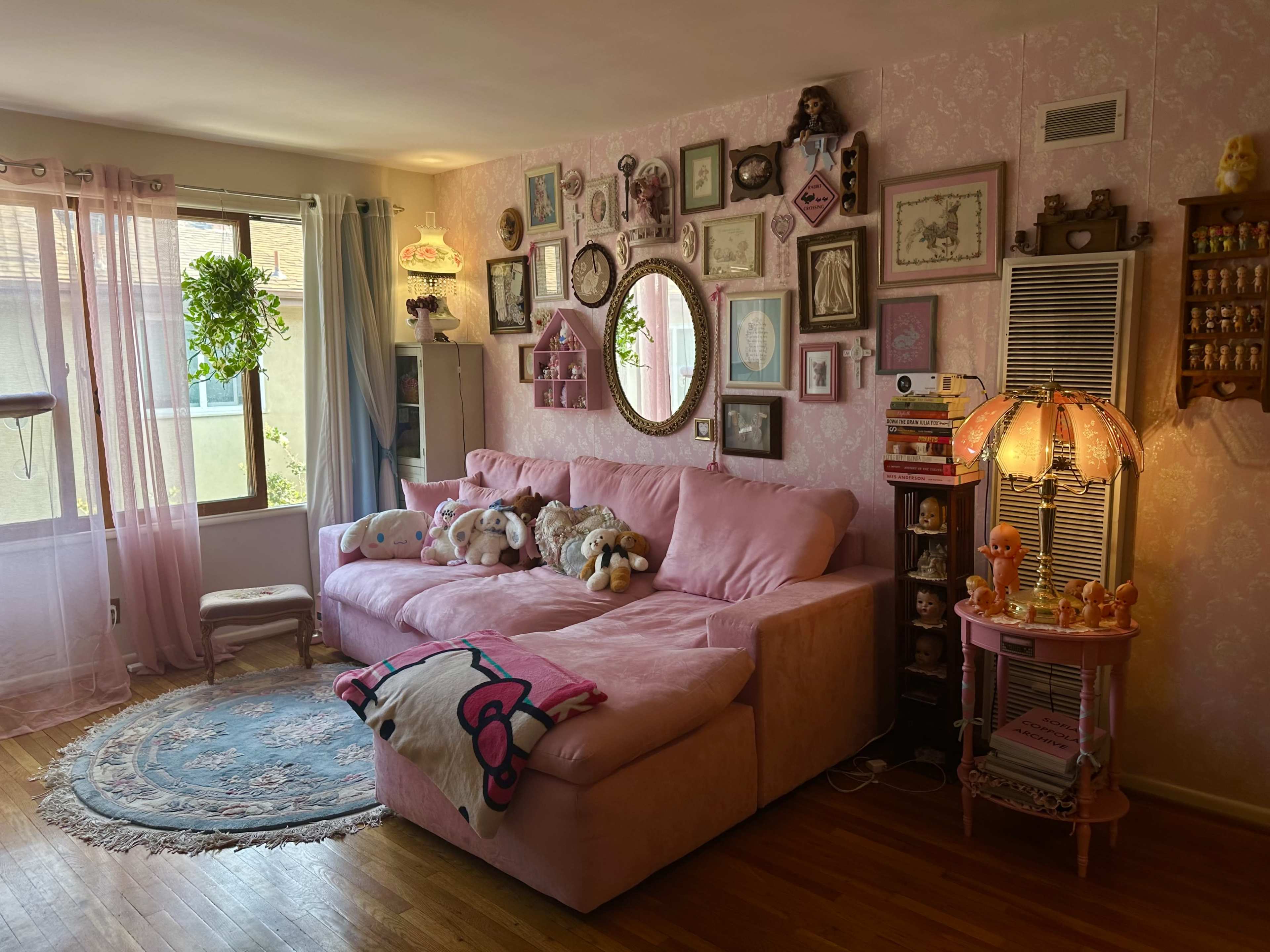 A cozy living room with a pink sectional sofa surrounded by framed artwork and decorative objects, including plants and a lamp.