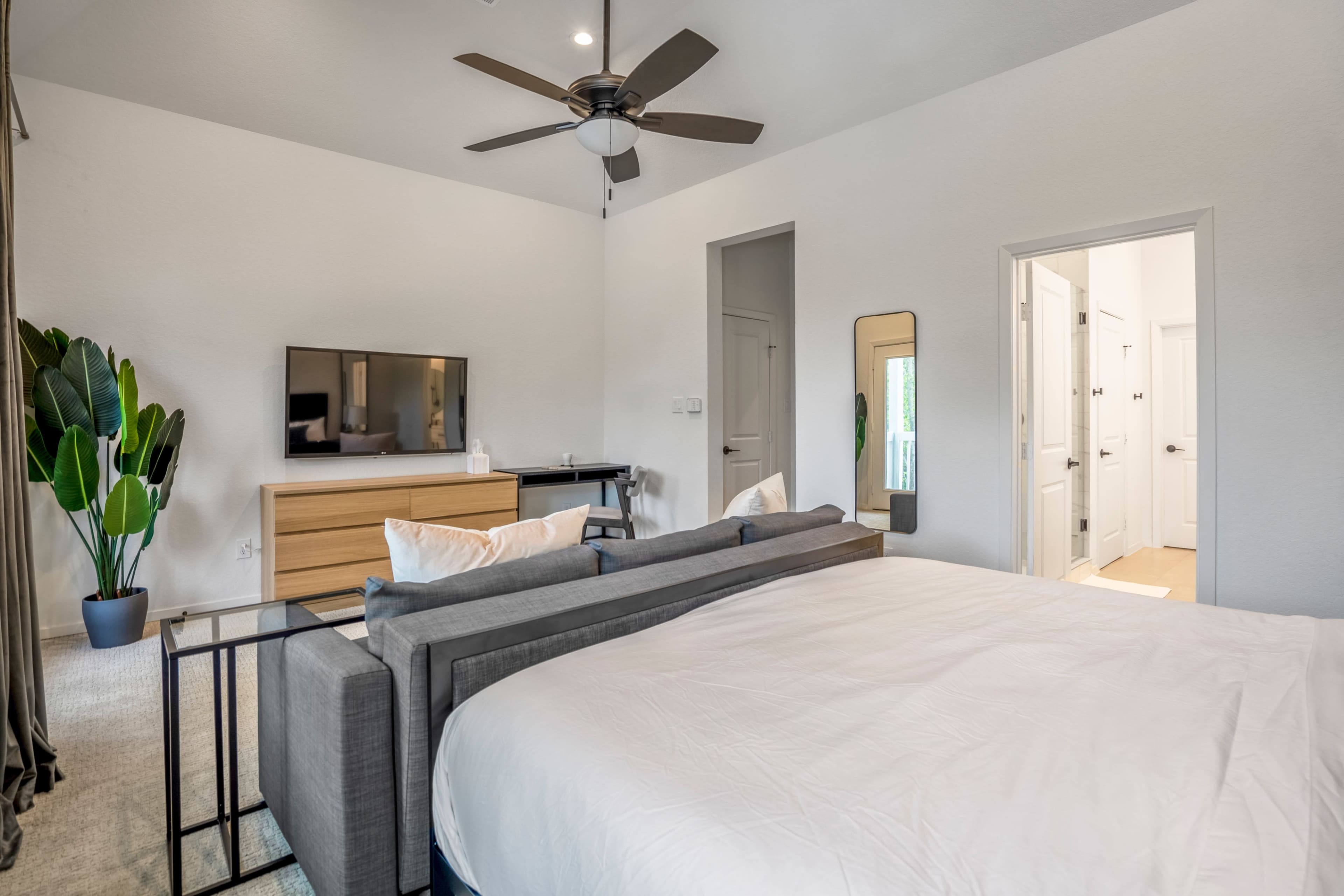 A modern bedroom featuring a bed, a dresser with a television, a standing mirror, and a potted plant next to a doorway.