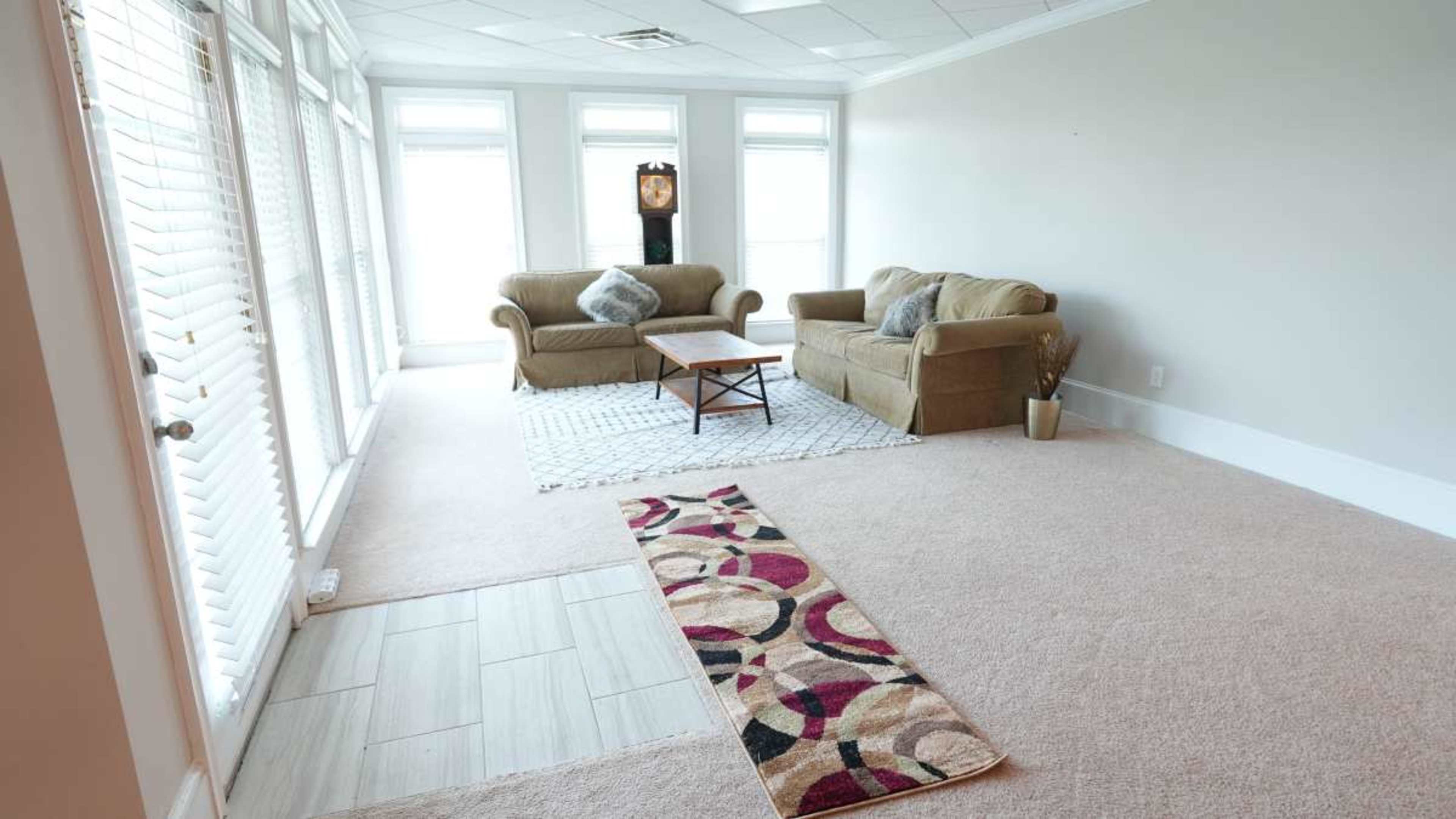 The image shows a spacious, well-lit living room furnished with two sofas and a coffee table, with large windows allowing natural light to enter.