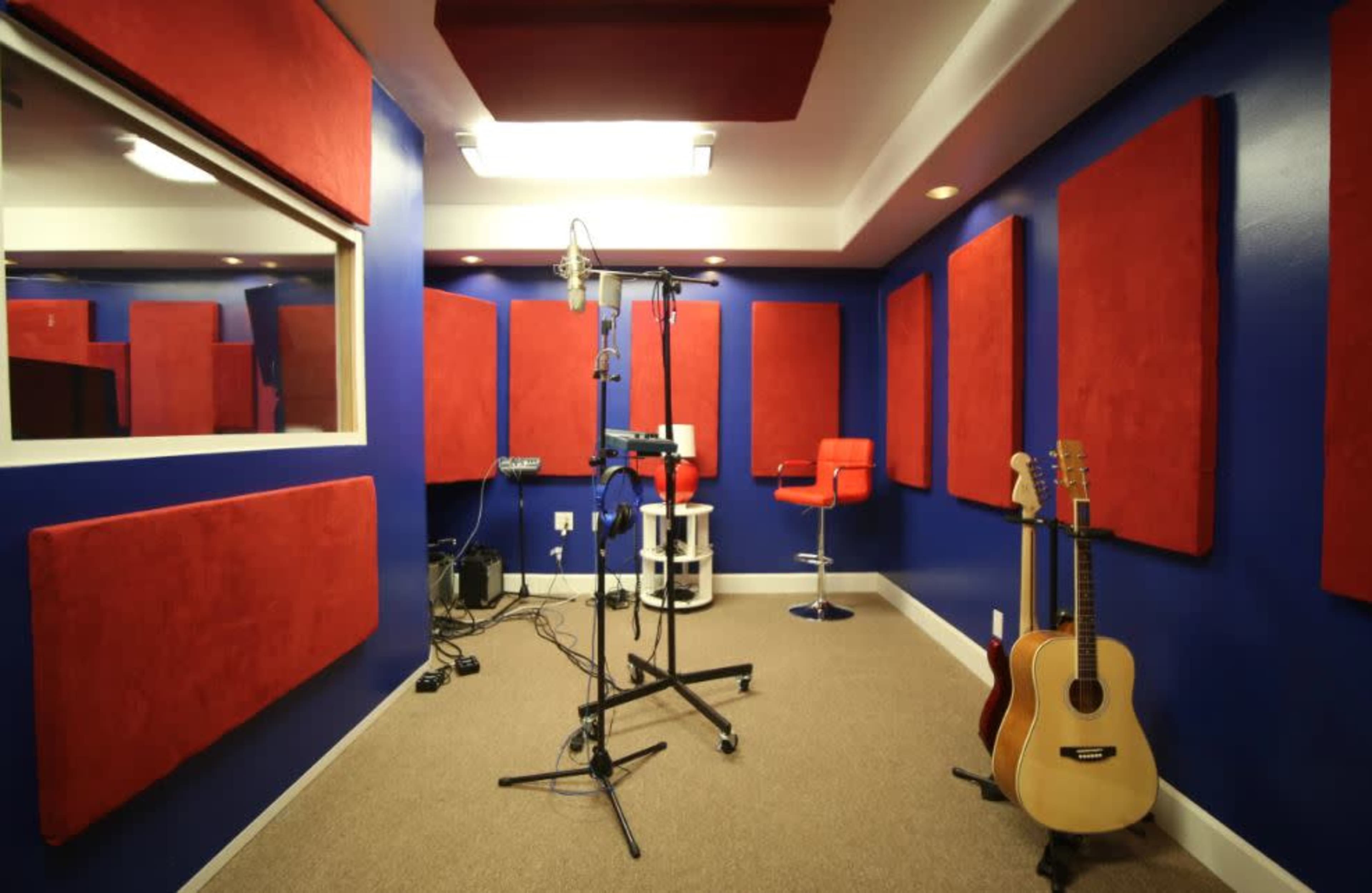 The image shows a soundproof recording studio featuring blue walls, red acoustic panels, a microphone stand, and guitars in the corner.