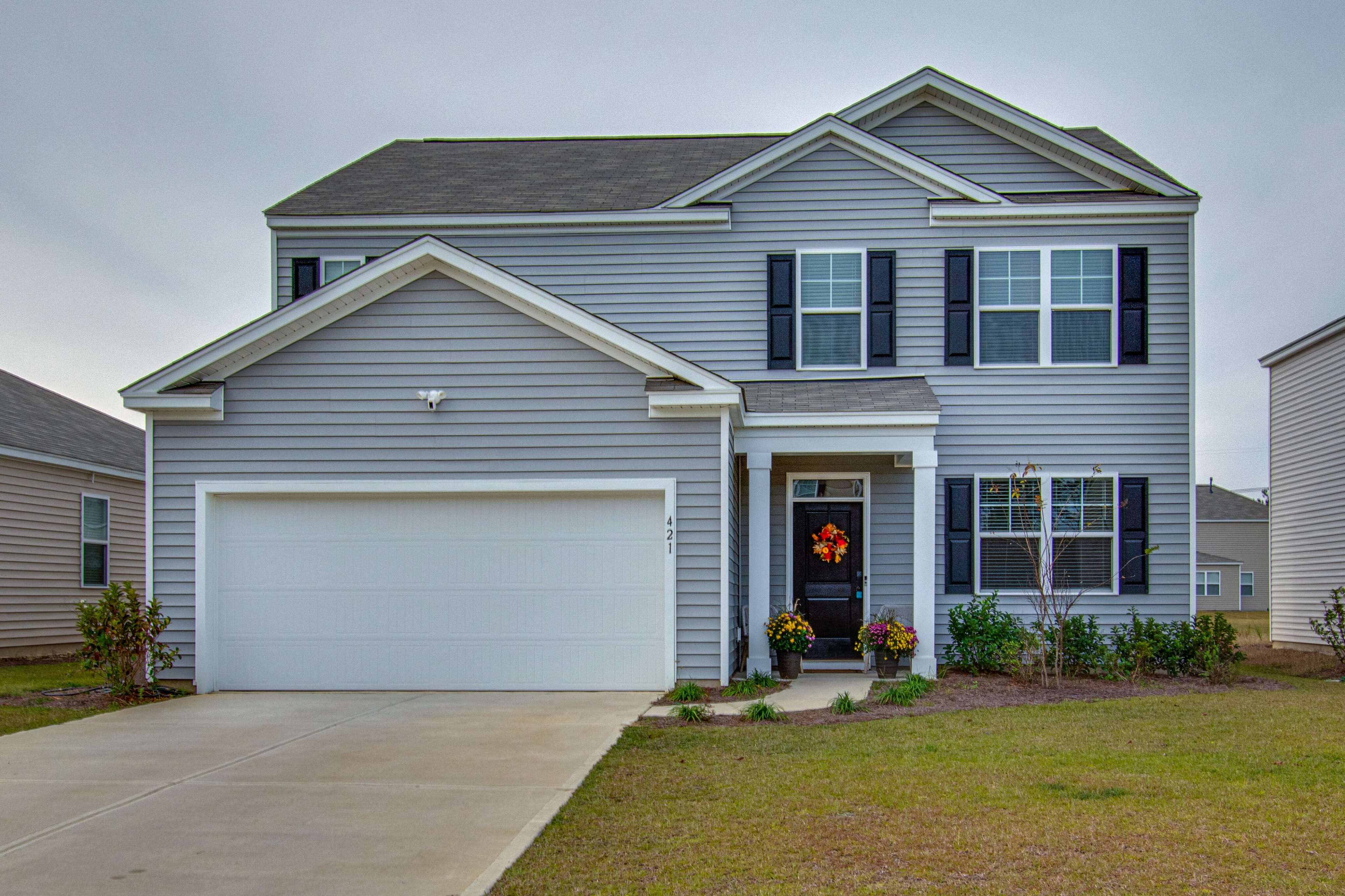 A two-story gray house with black shutters and a white garage door features a front porch adorned with a wreath and flower beds on either side.