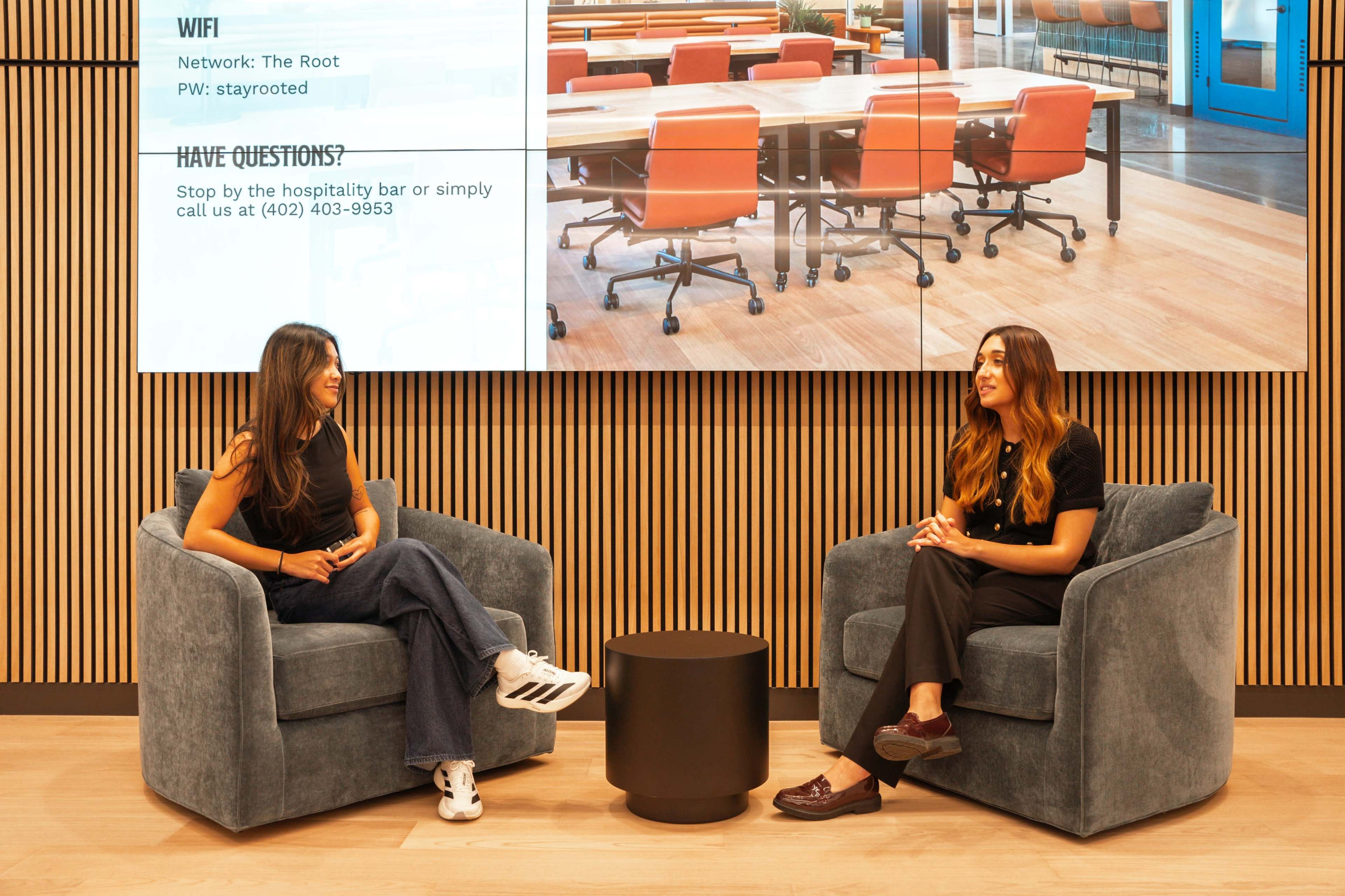 Two women sit in cozy blue armchairs, engaged in conversation, with a digital display behind them showing WiFi information and a hospitality bar contact number.
