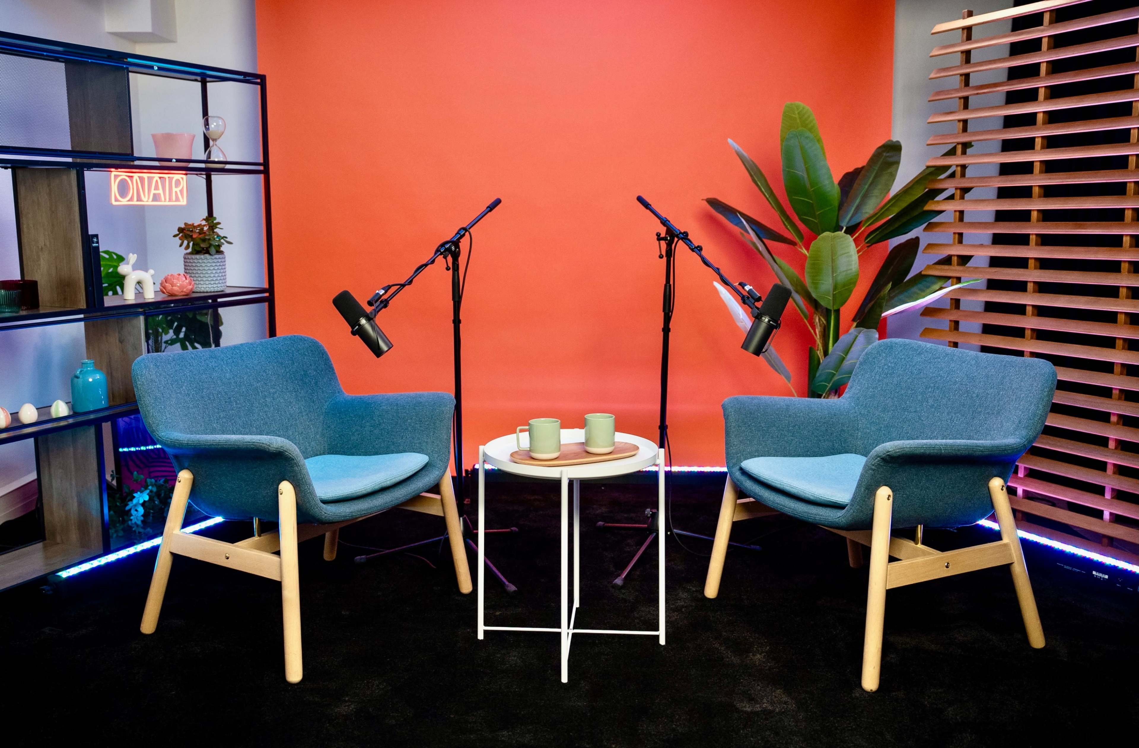 The image features two blue armchairs set around a small white table, with two microphones positioned nearby against an orange backdrop and a potted plant in the scene.