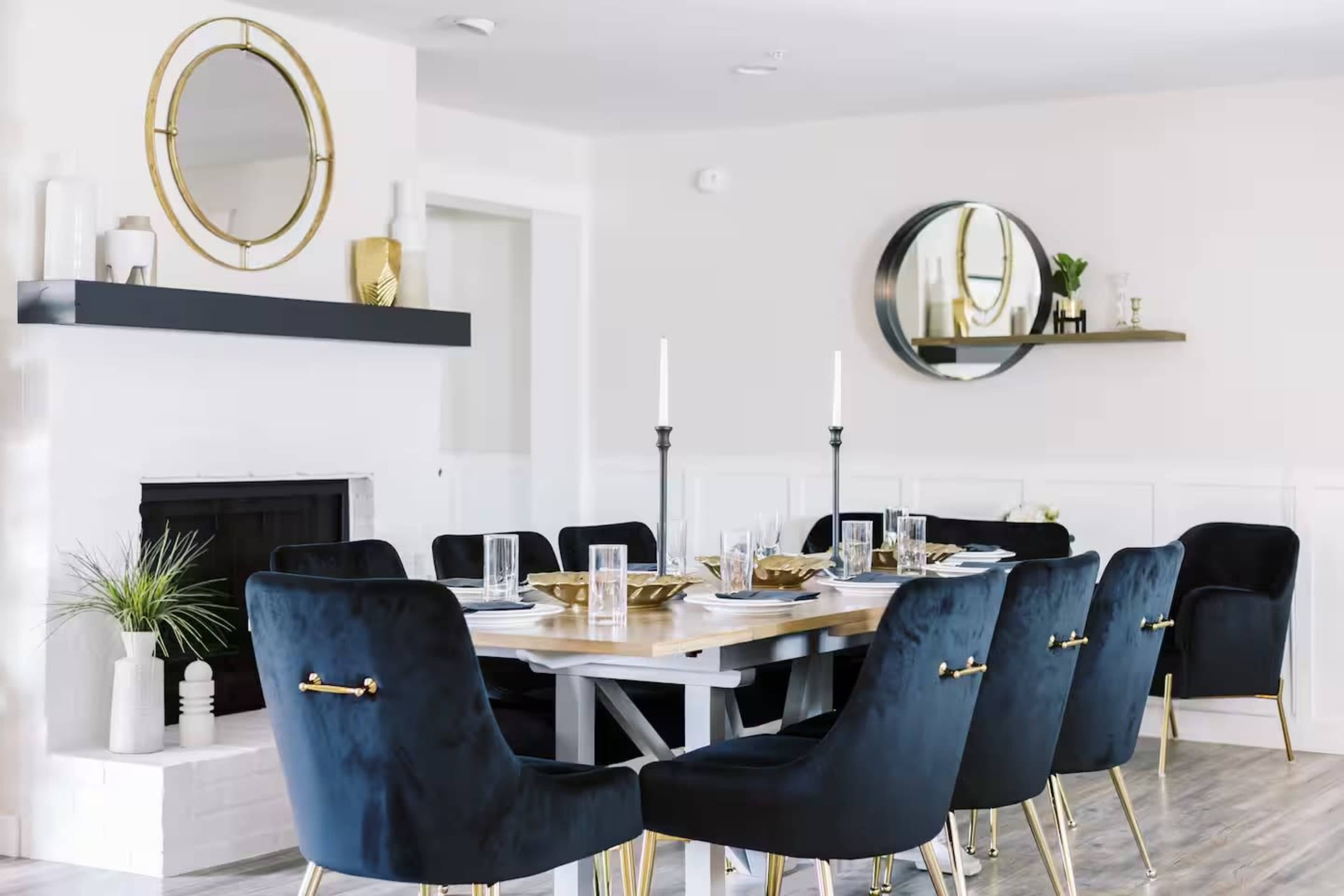 The image shows a modern dining room with a long table set for a meal, surrounded by dark blue chairs and featuring a minimalist fireplace and decorative mirrors on the walls.