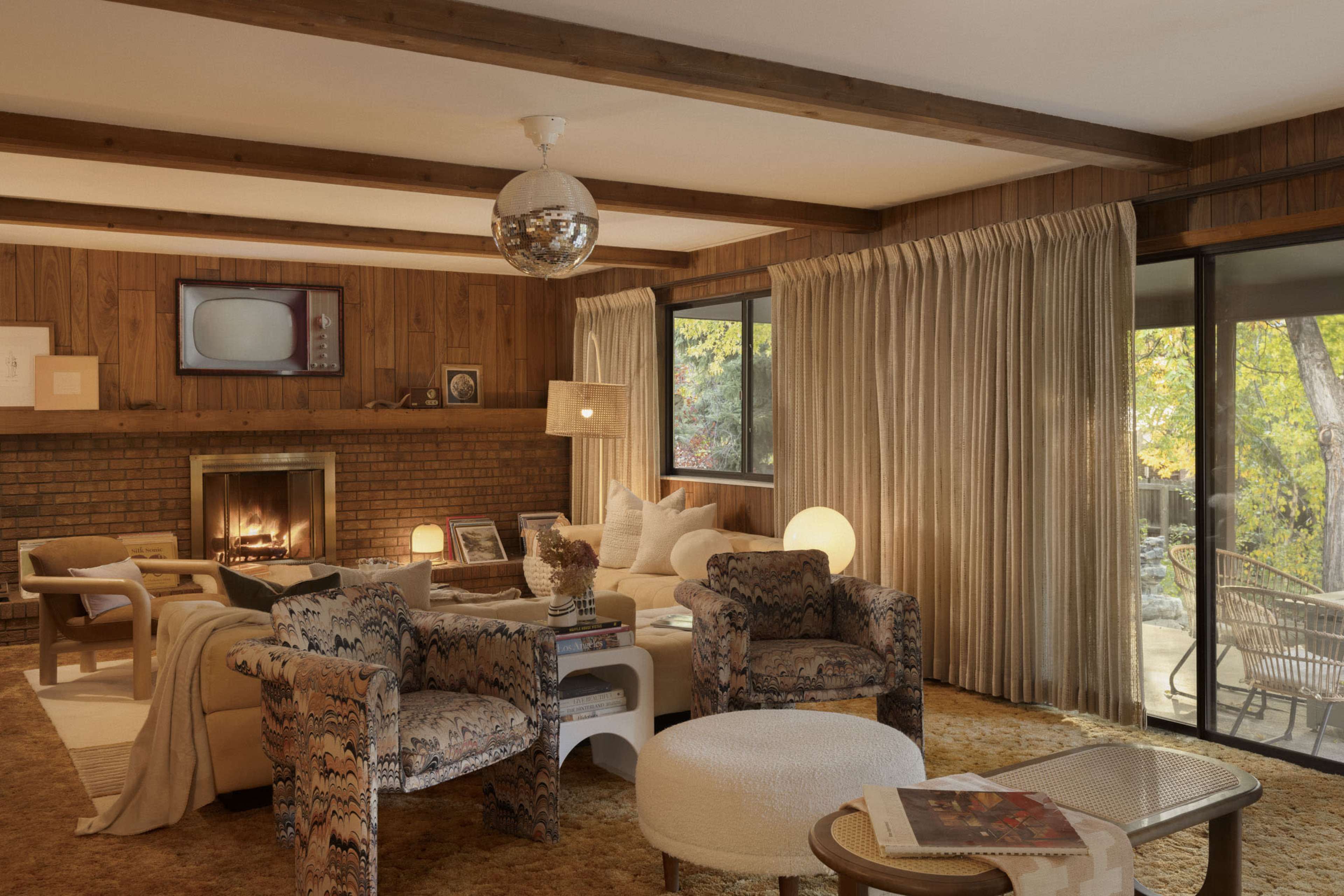 The living room features wooden beams, a brick fireplace, and seating with patterned chairs, illuminated by natural light through large windows.