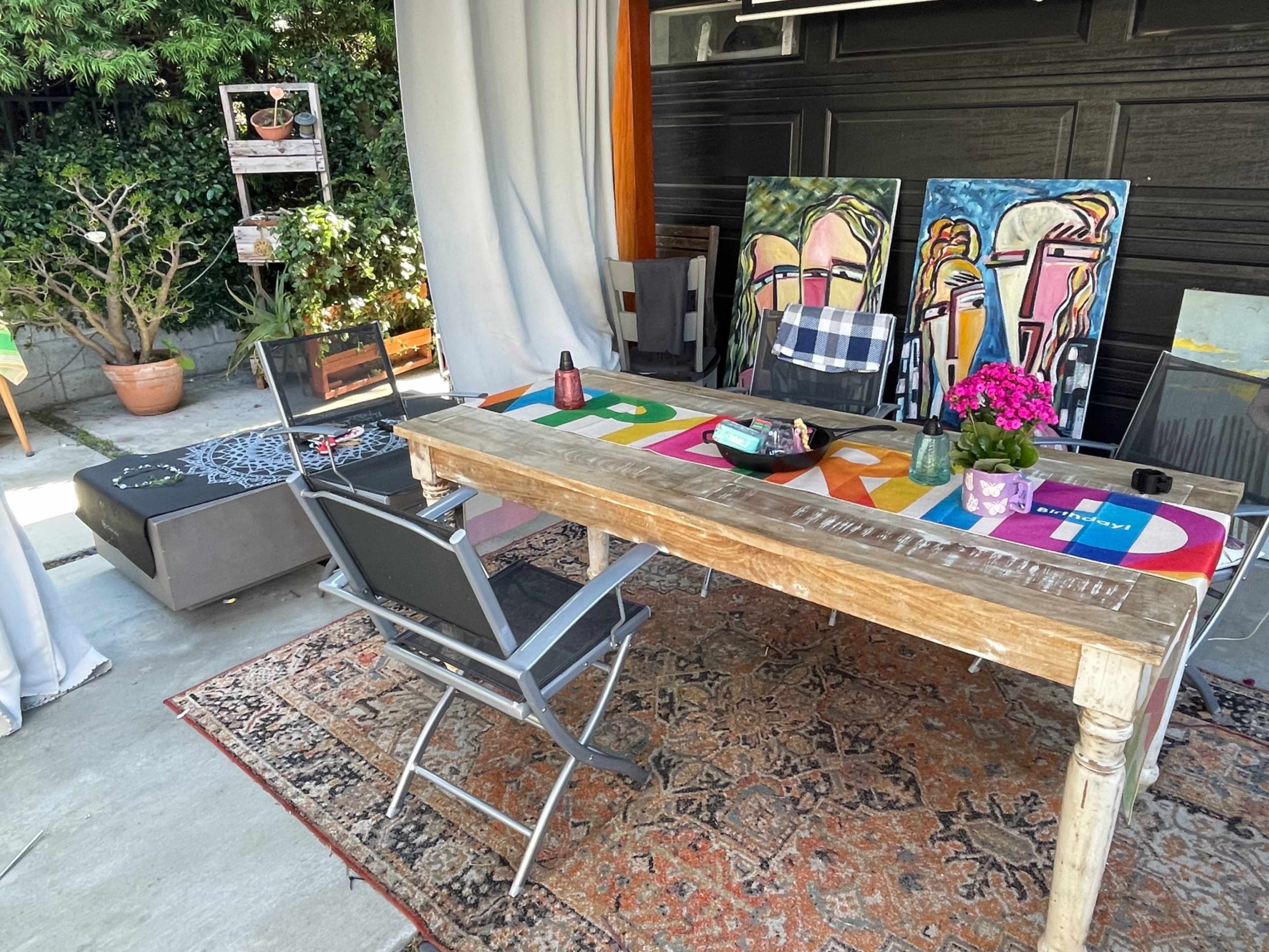 An outdoor seating area features a wooden table with colorful artwork on display, surrounded by chairs and potted plants.