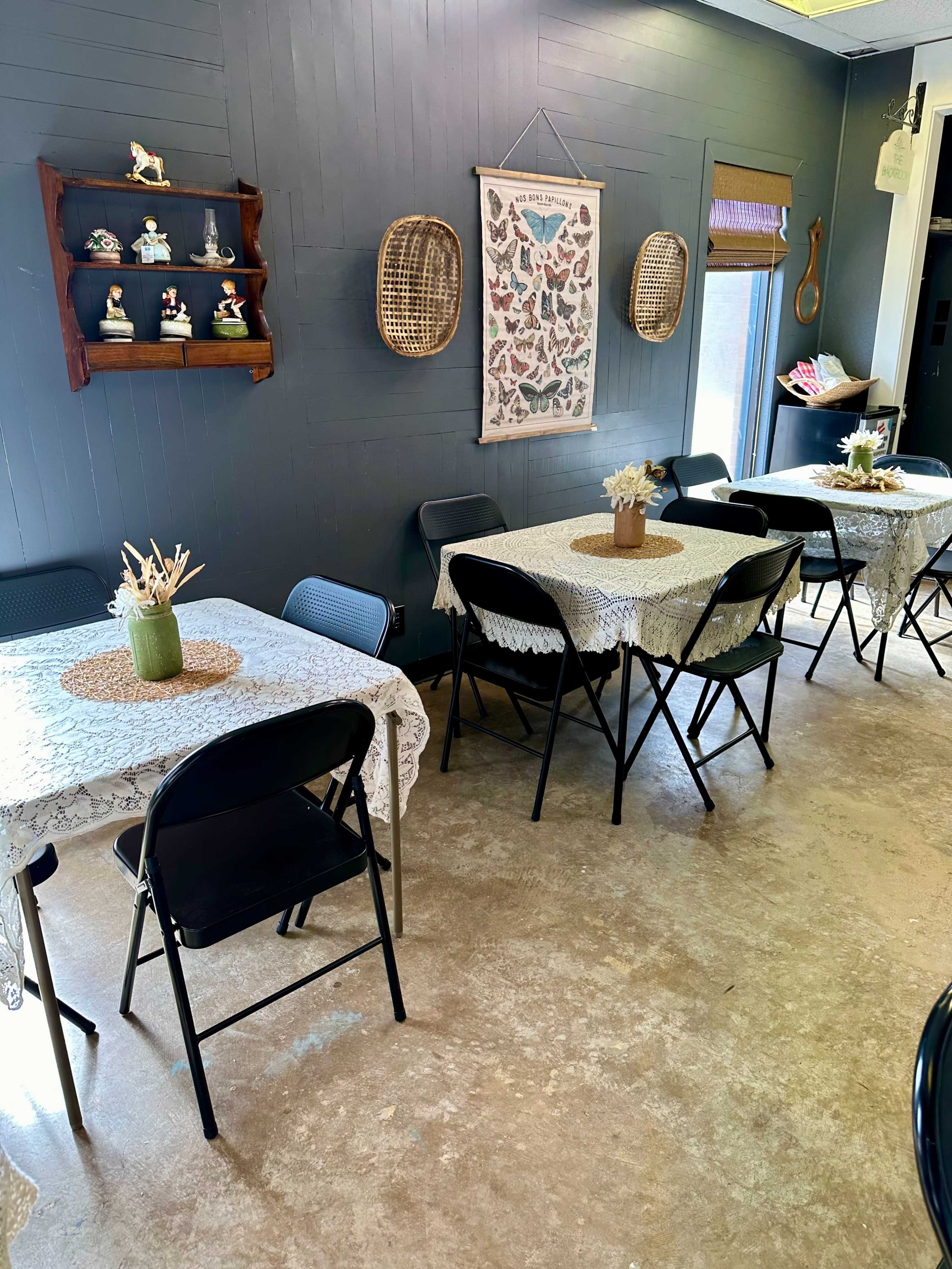 A small dining area with several tables covered in white lace tablecloths, adorned with floral centerpieces, against a backdrop of dark gray walls and decorative wall hangings.