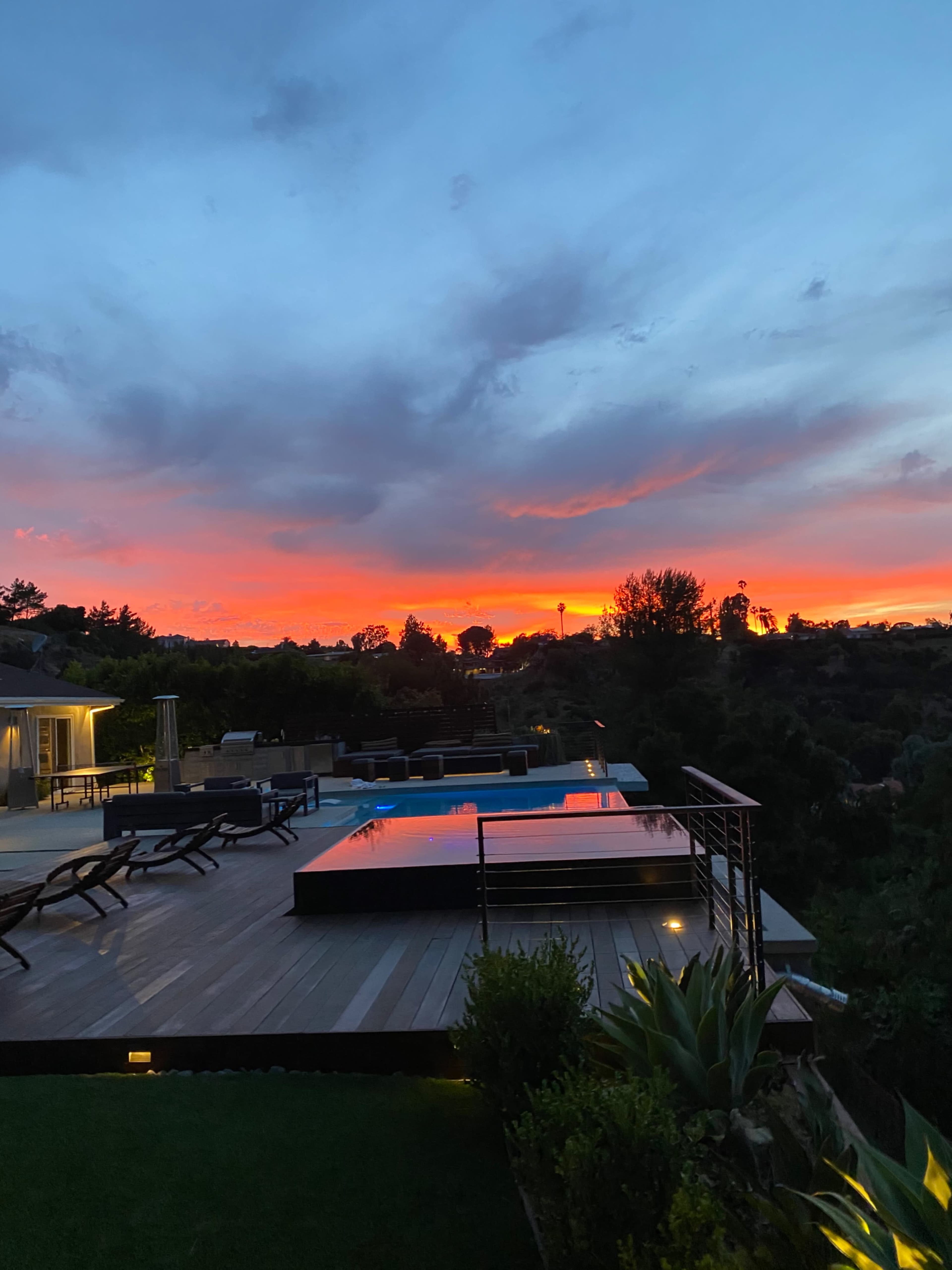 A modern deck featuring an infinity pool overlooks a vibrant sunset sky with clouds.