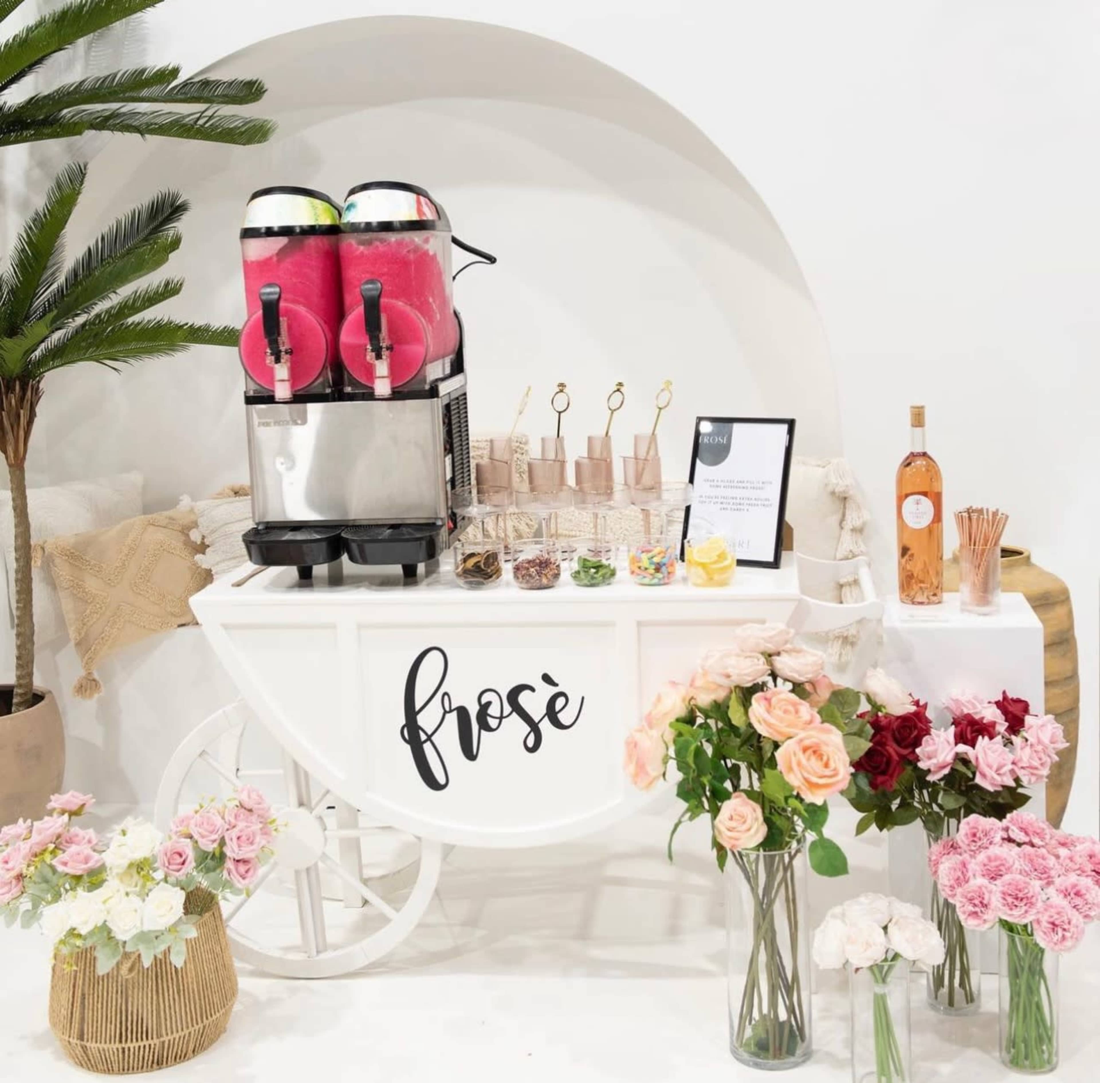 A frose cart featuring two beverage dispensers, glassware, and a selection of toppings, surrounded by various floral arrangements.