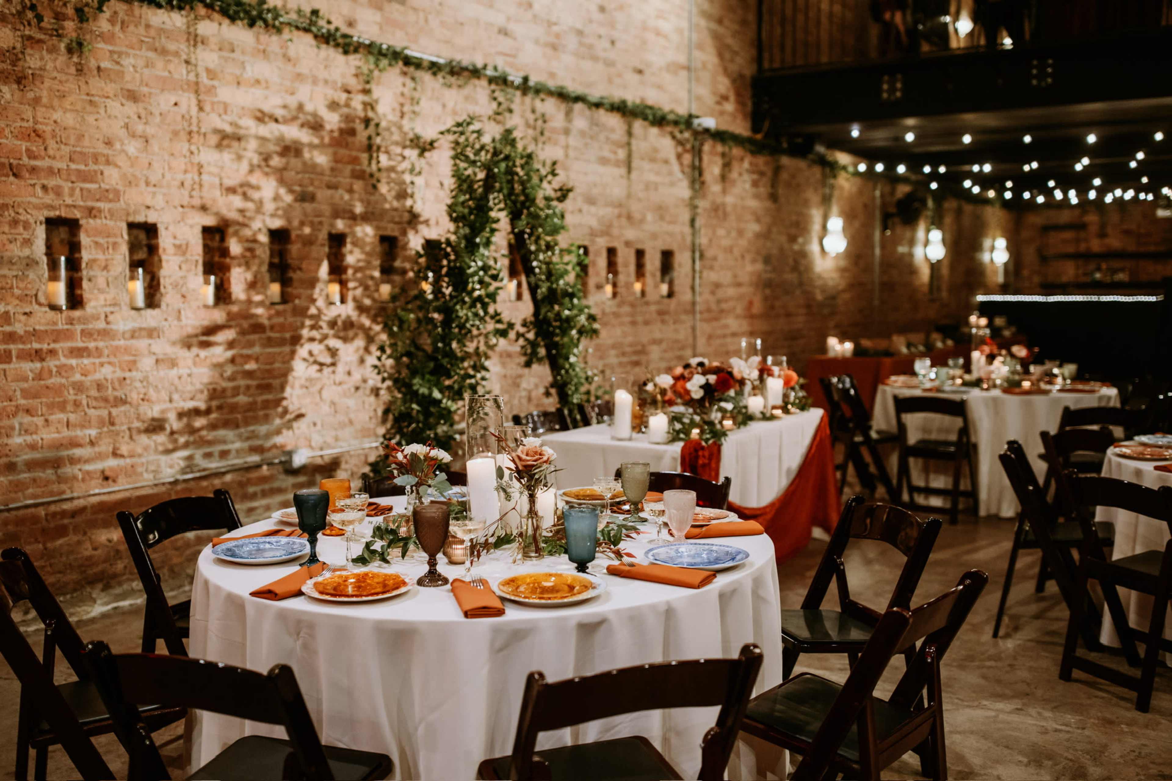 A rustic event space features elegantly set tables with colorful plates, candles, and greenery against a brick wall backdrop.