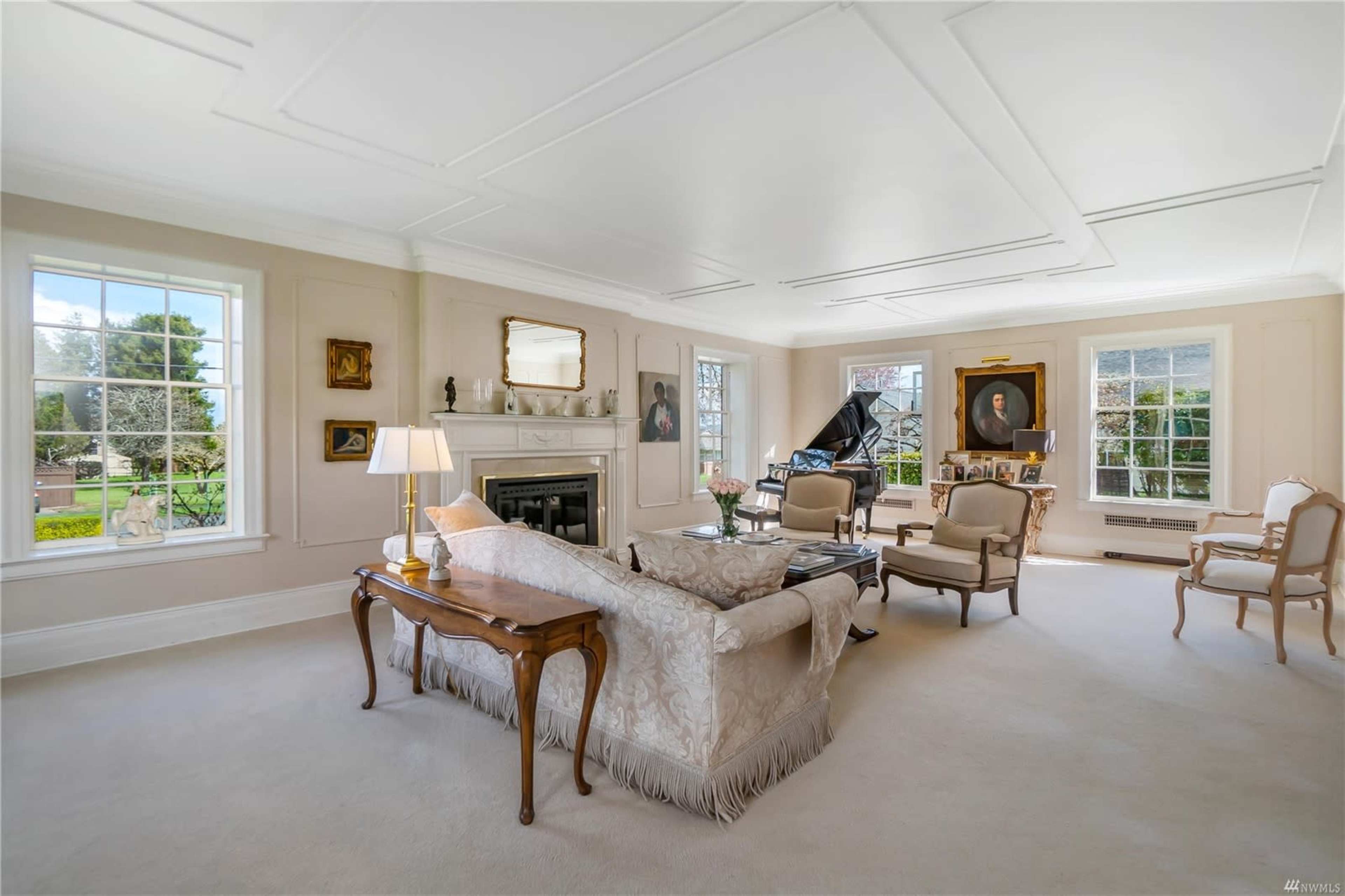 The spacious living room features a fireplace, large windows, a grand piano, and elegant furniture arranged around a central seating area.
