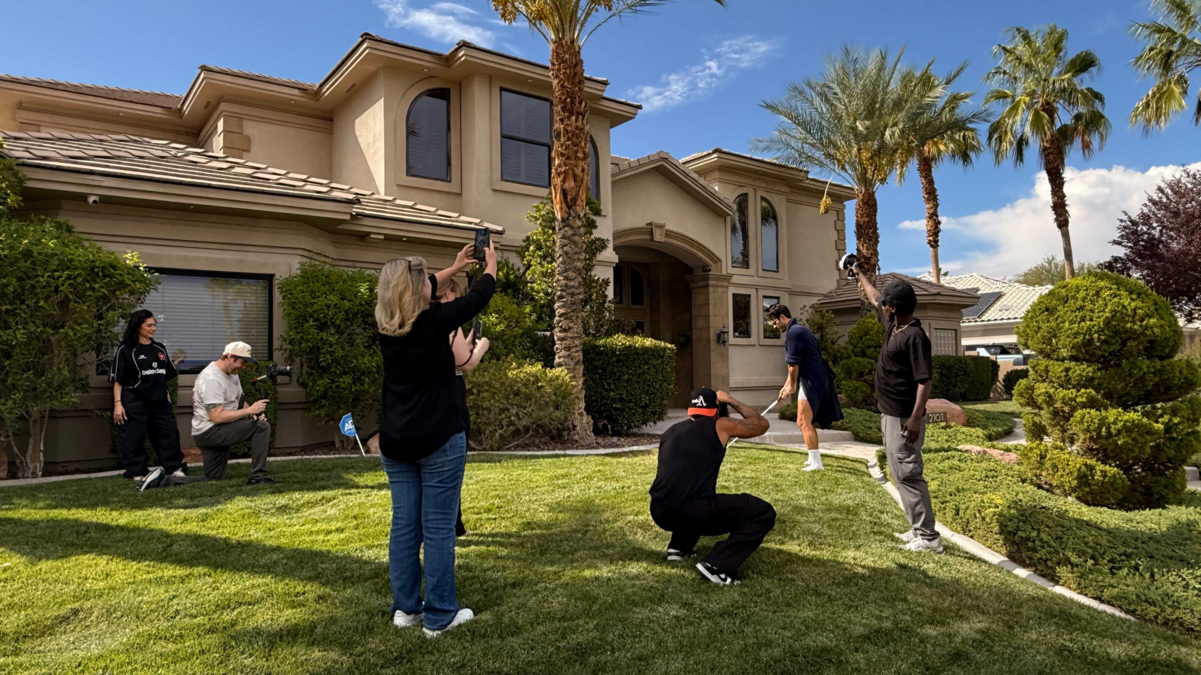 A group of six individuals is gathered in the front yard of a house with palm trees, capturing photos with cameras and smartphones.