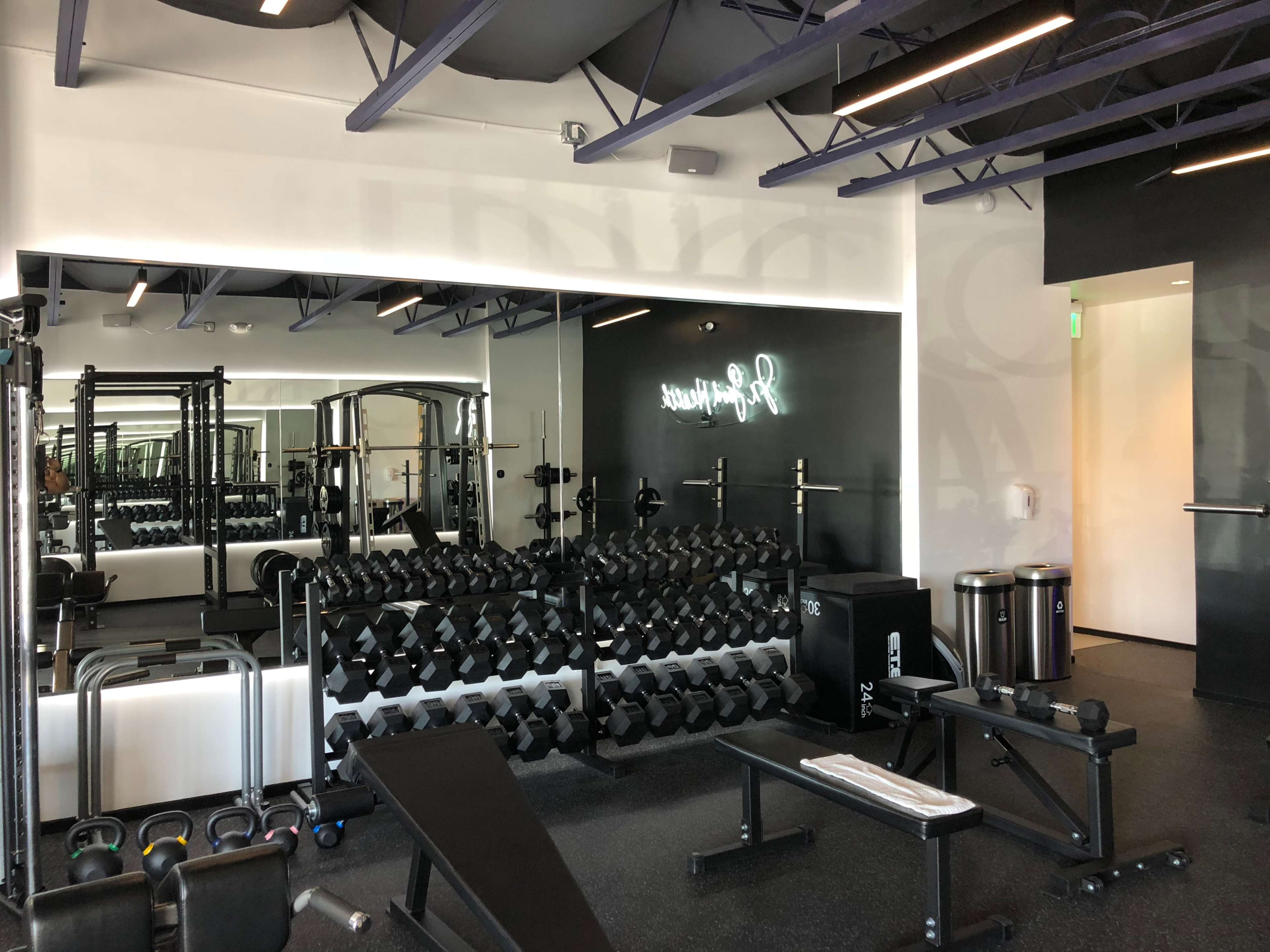 A modern gym interior with a weightlifting area featuring a rack of dumbbells, benches, and various exercise equipment.