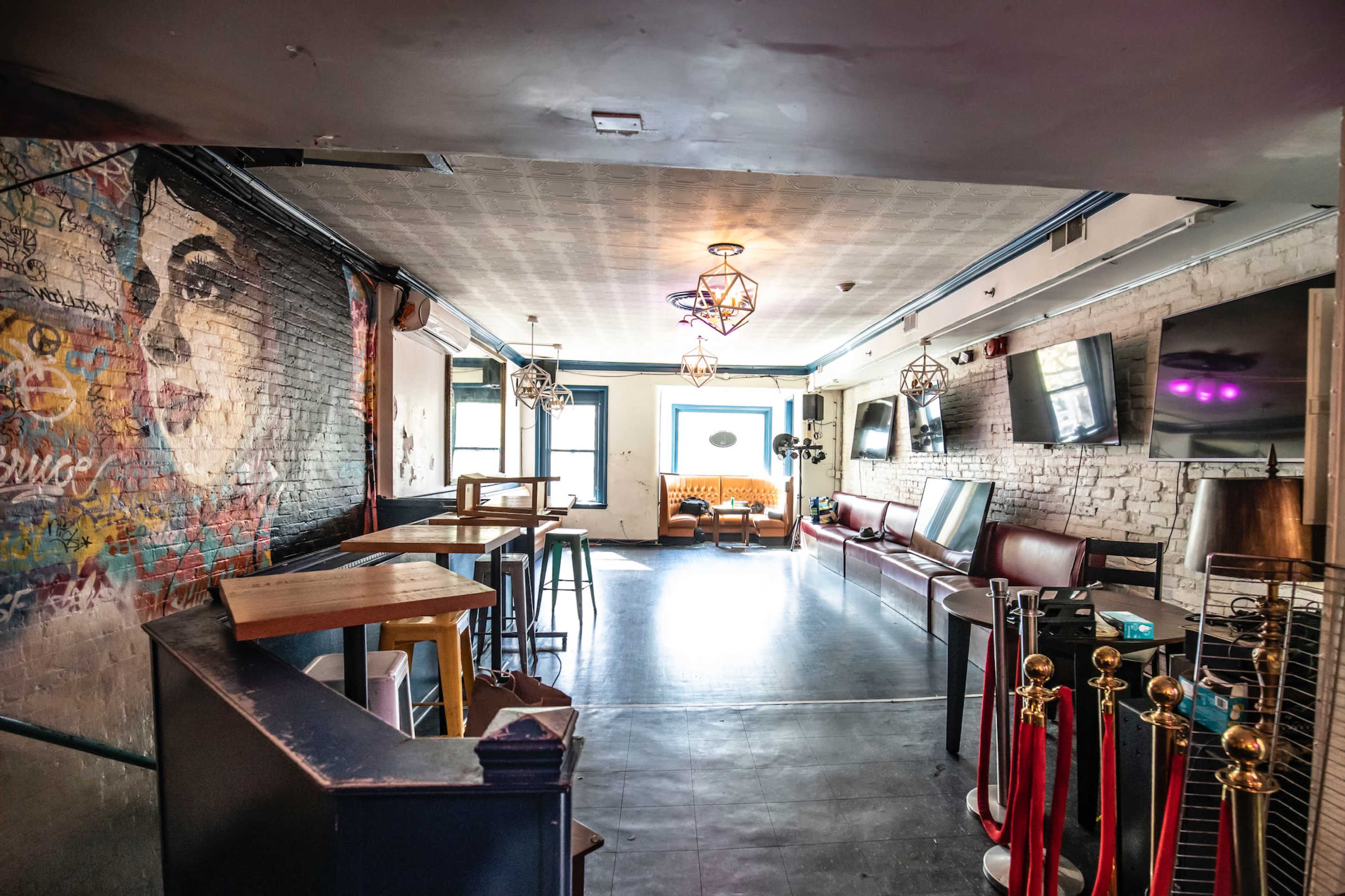 The image shows the interior of a bar or lounge featuring a mural on one wall, seating areas, and decorative lighting.
