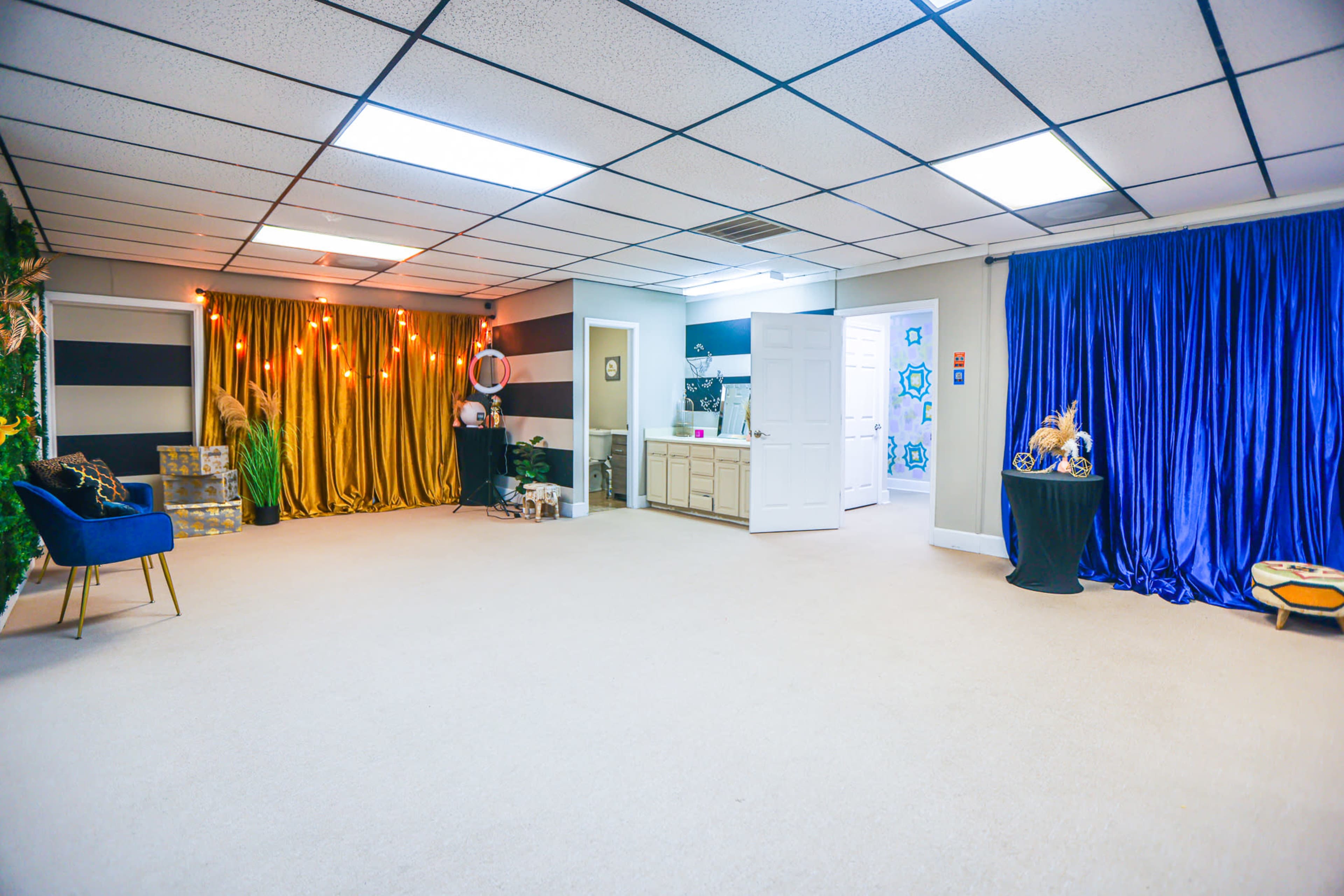The image shows a spacious room with beige carpet, featuring colorful curtains in gold and blue, a lounge chair, and various decorative items, along with doors leading to other areas.