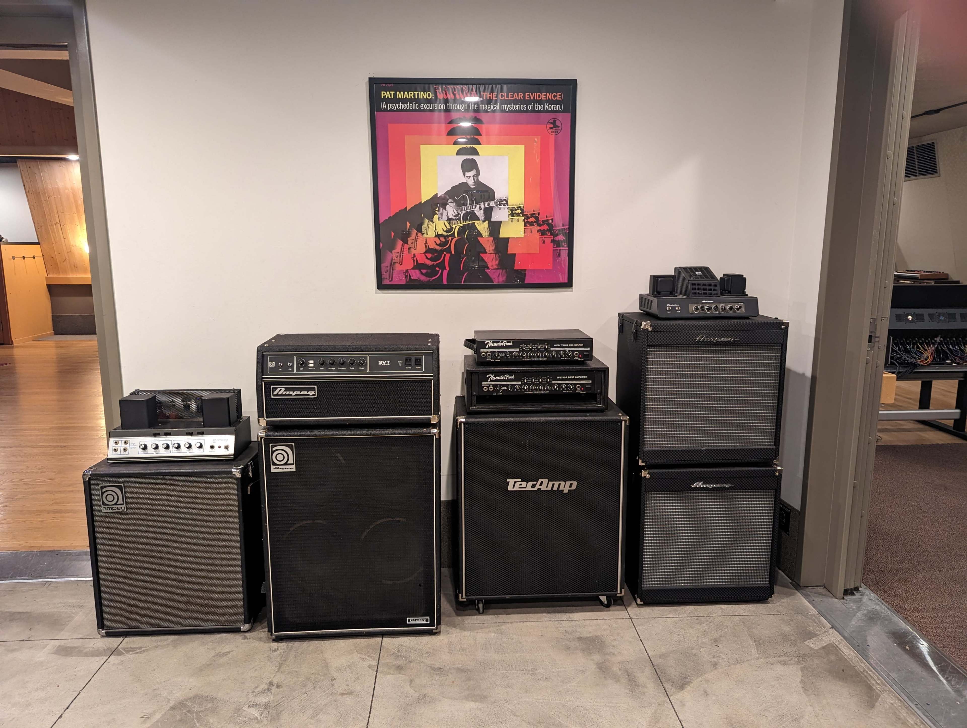 Several amplifiers stacked against a wall, with a framed poster of a musician above them.