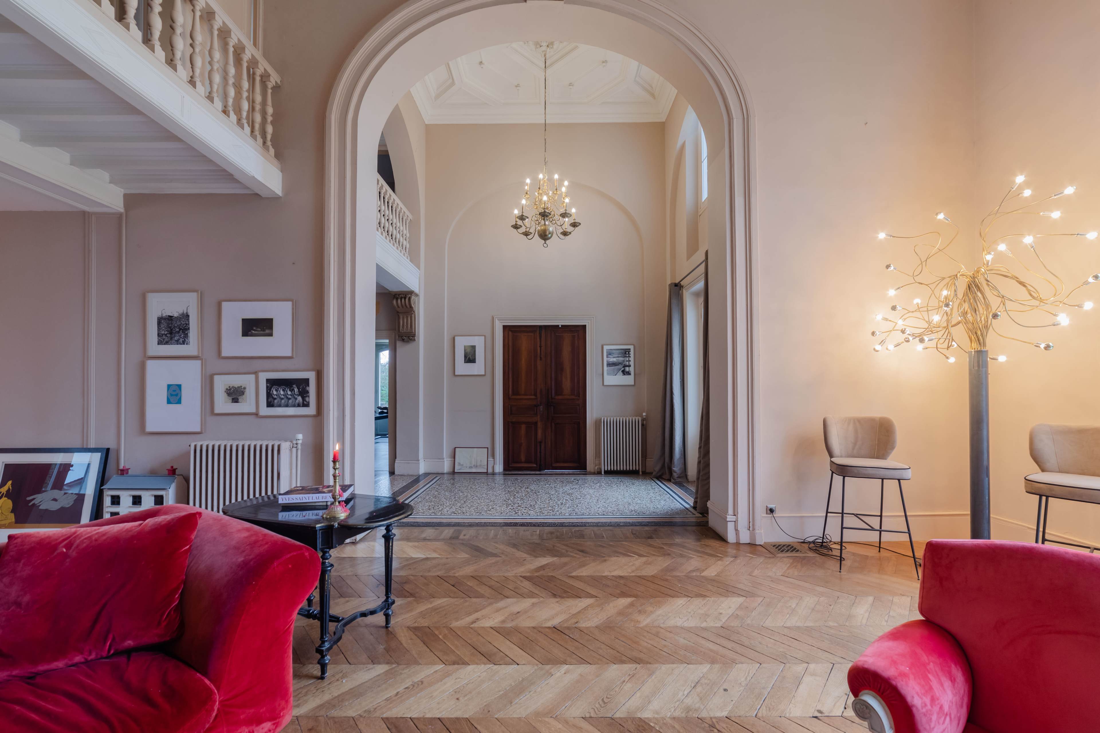 A spacious interior with wooden flooring, featuring a large archway, a chandelier, and a modern light fixture beside two red sofas.