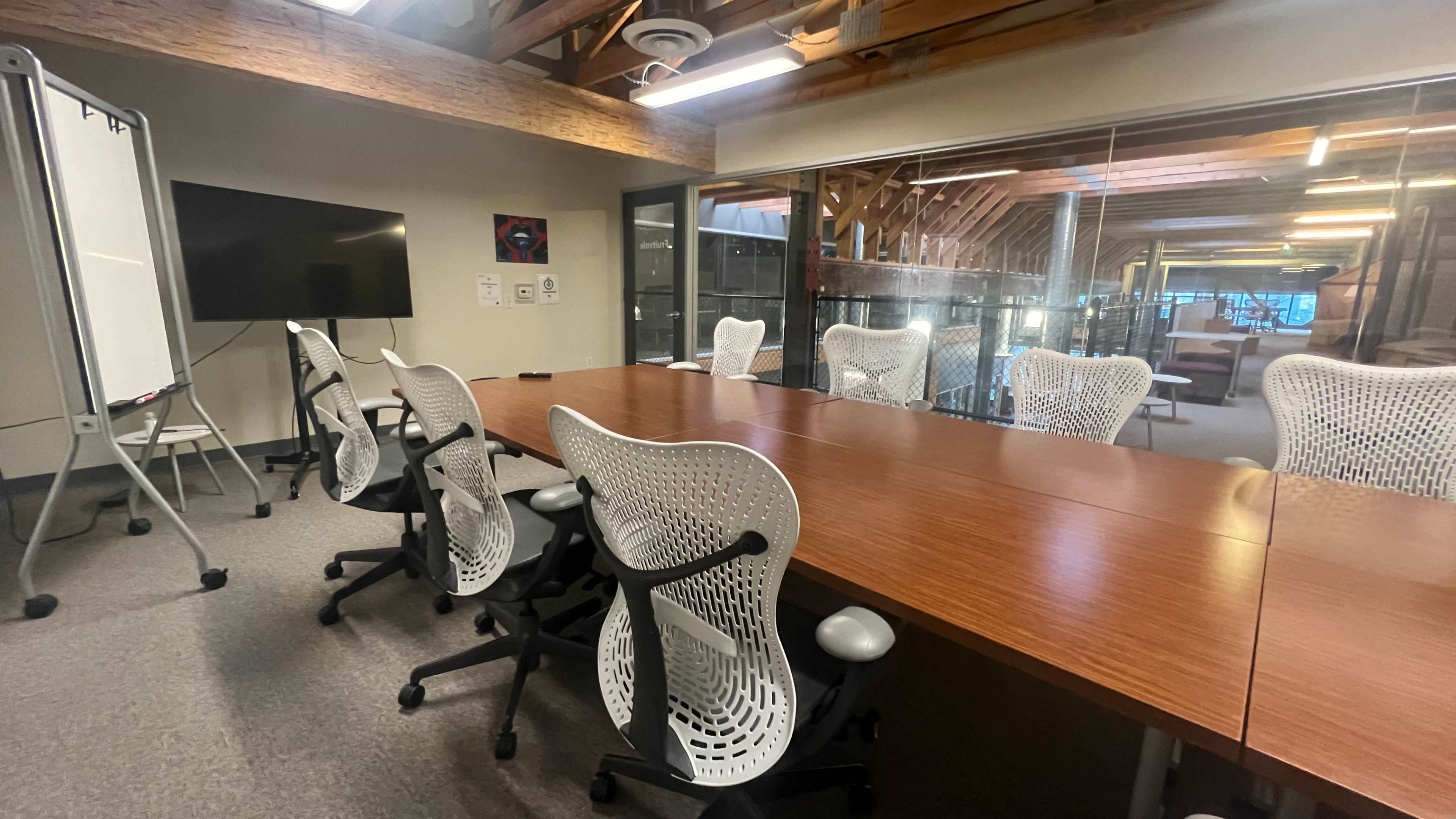 A conference room features a long wooden table surrounded by modern white office chairs and a large screen at one end.