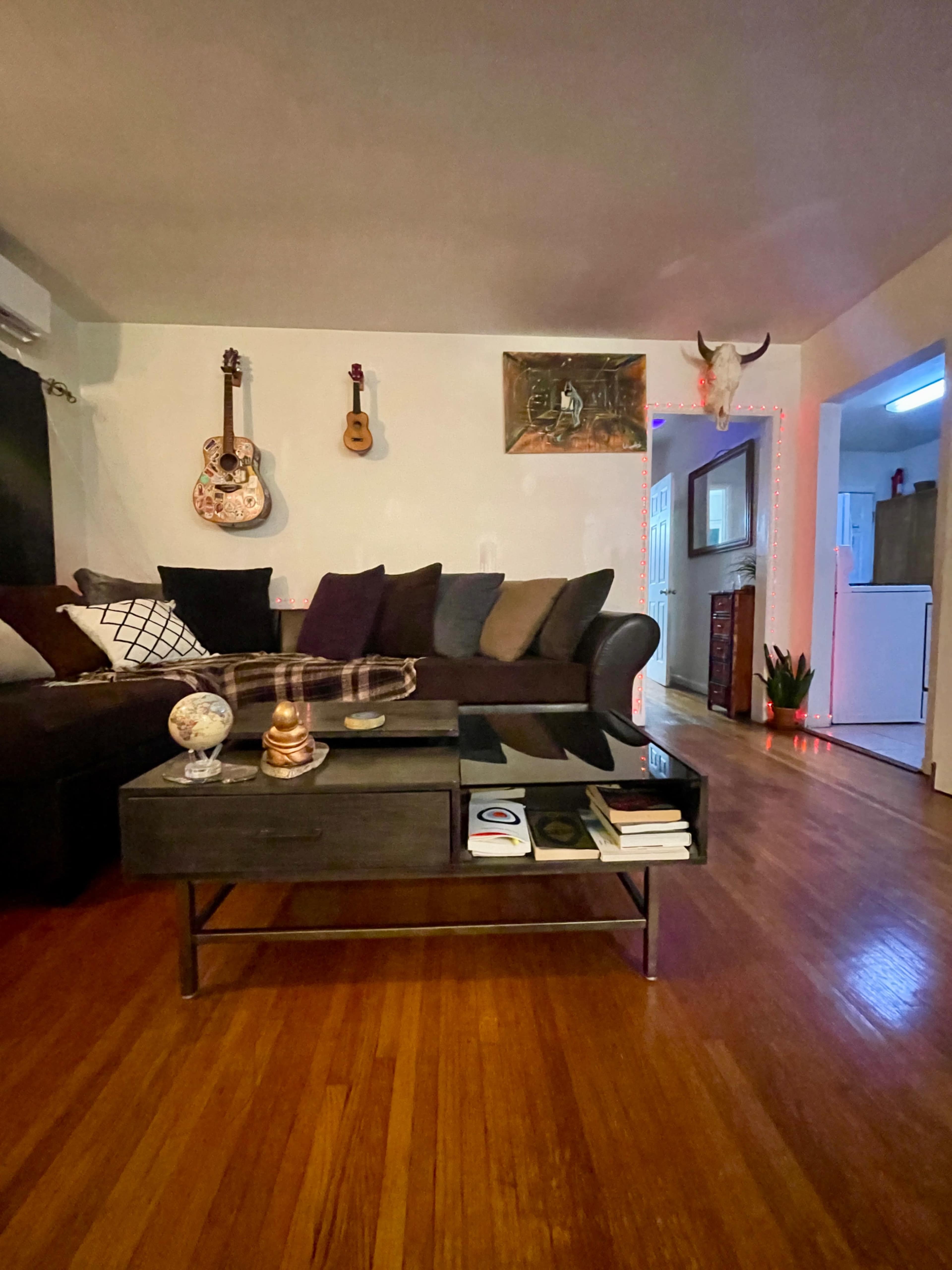 The living room features a dark sofa with various cushions, a glass coffee table, and wall-mounted guitars.