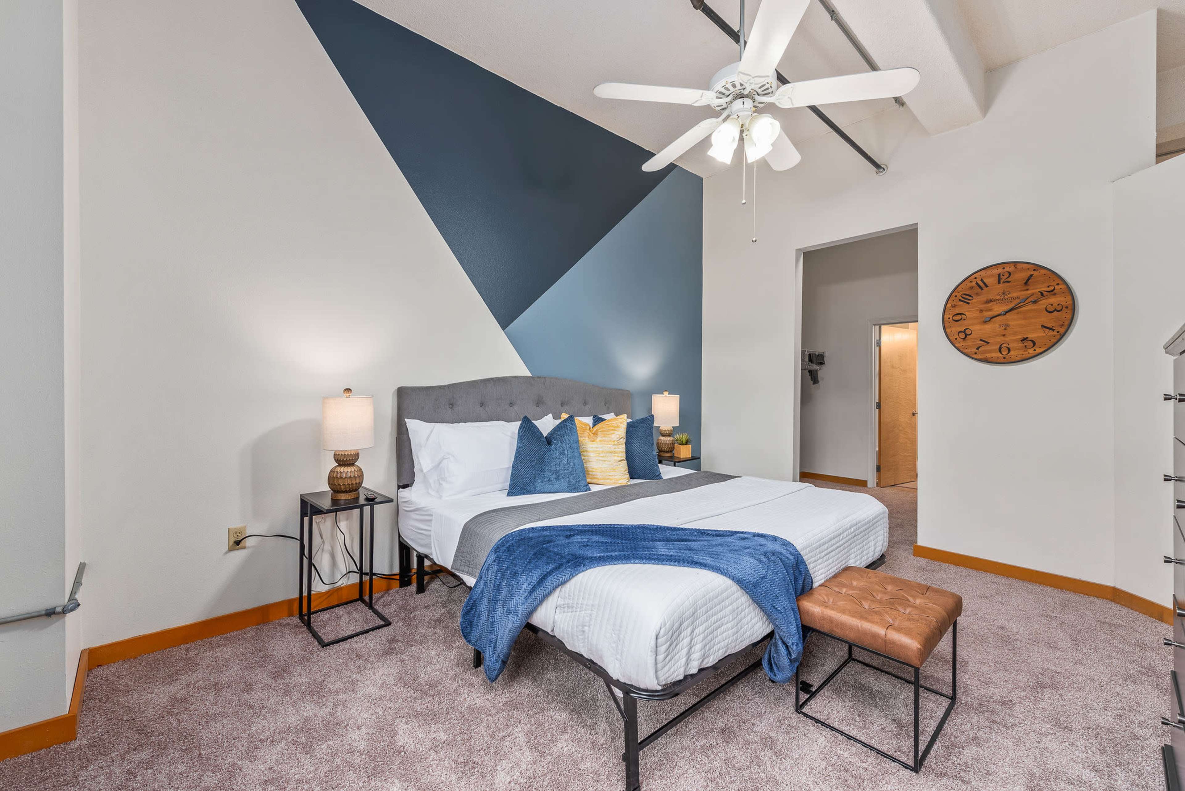 A neatly arranged bedroom features a bed with a gray headboard, decorative pillows, a blue throw blanket, and a wall painted with geometric shapes.