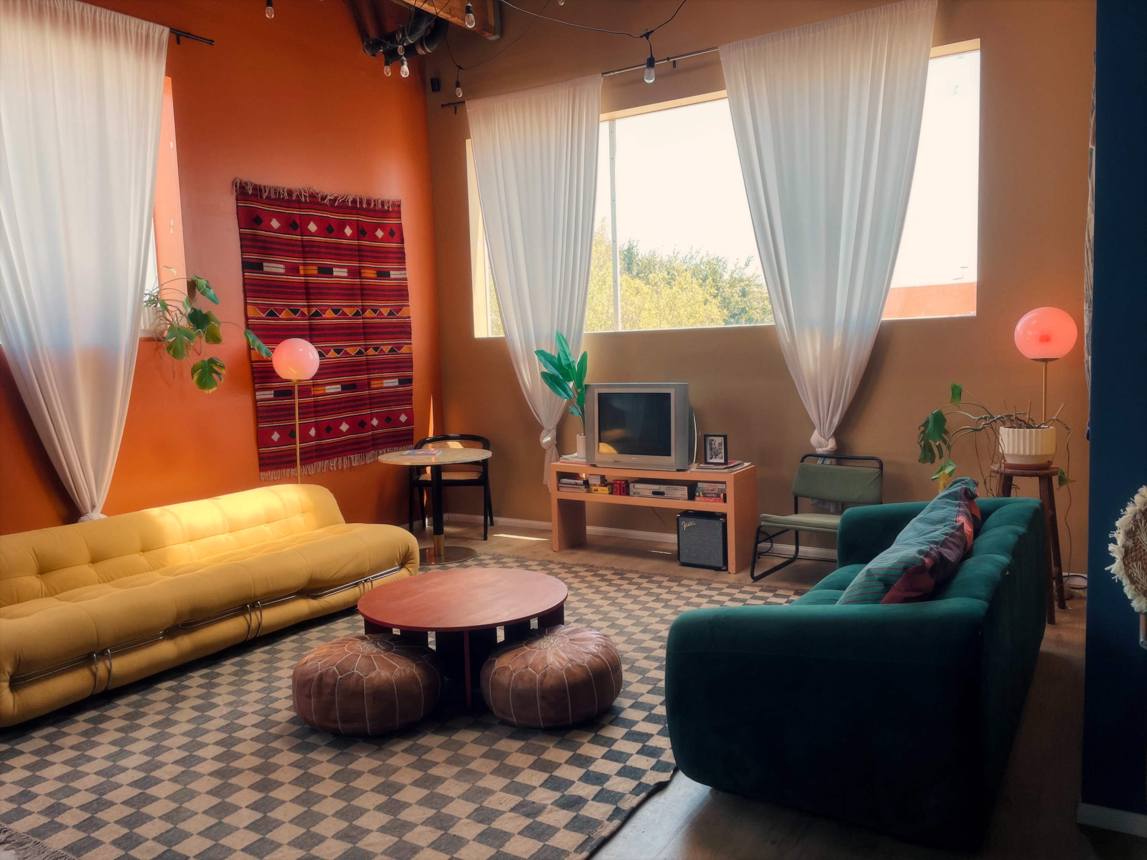 The image shows a cozy living room with two sofas, a round coffee table, plants, and large windows draped with white curtains.