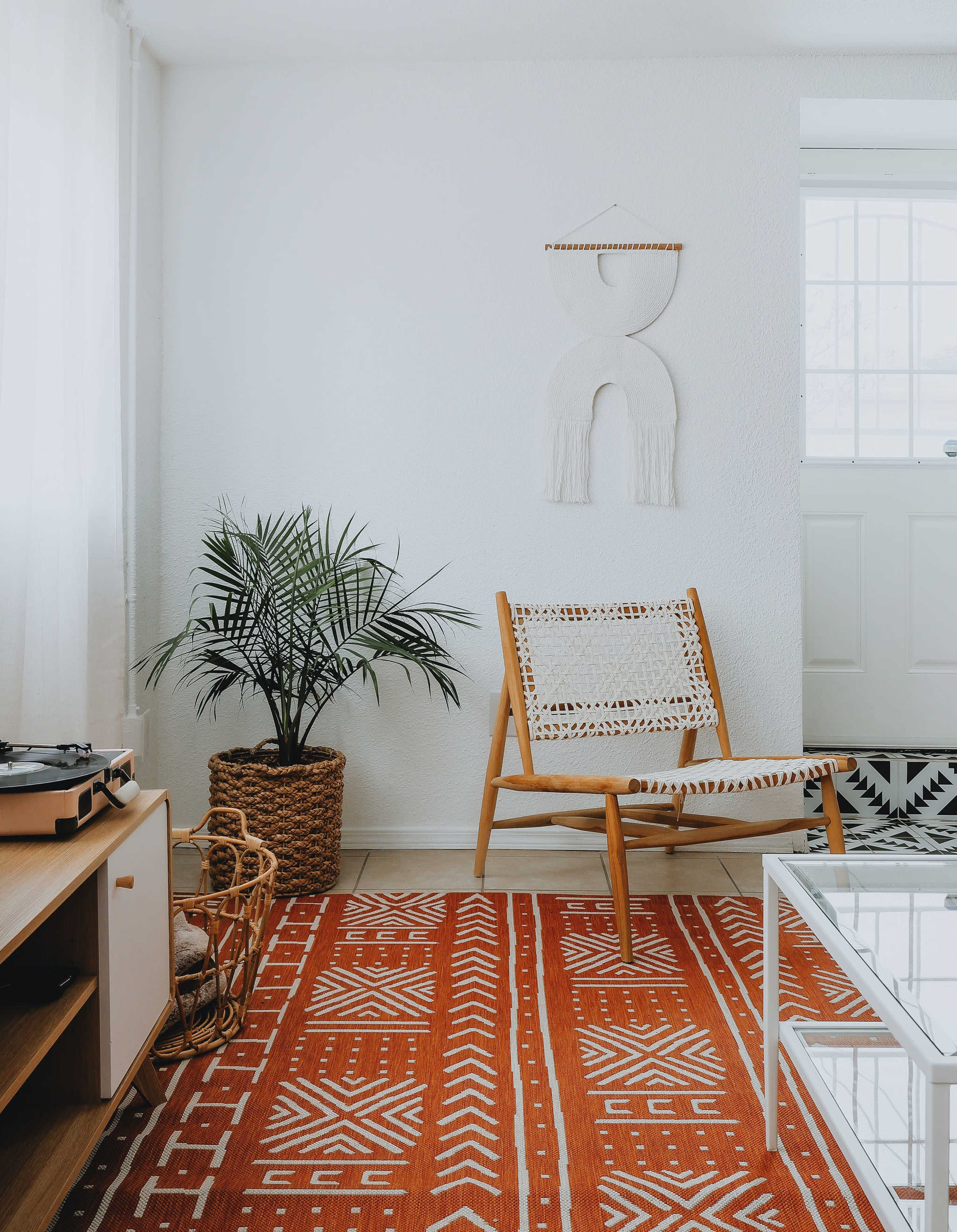 A cozy living room features a patterned rug, a wooden chair with a woven seat, a potted plant, and a record player.