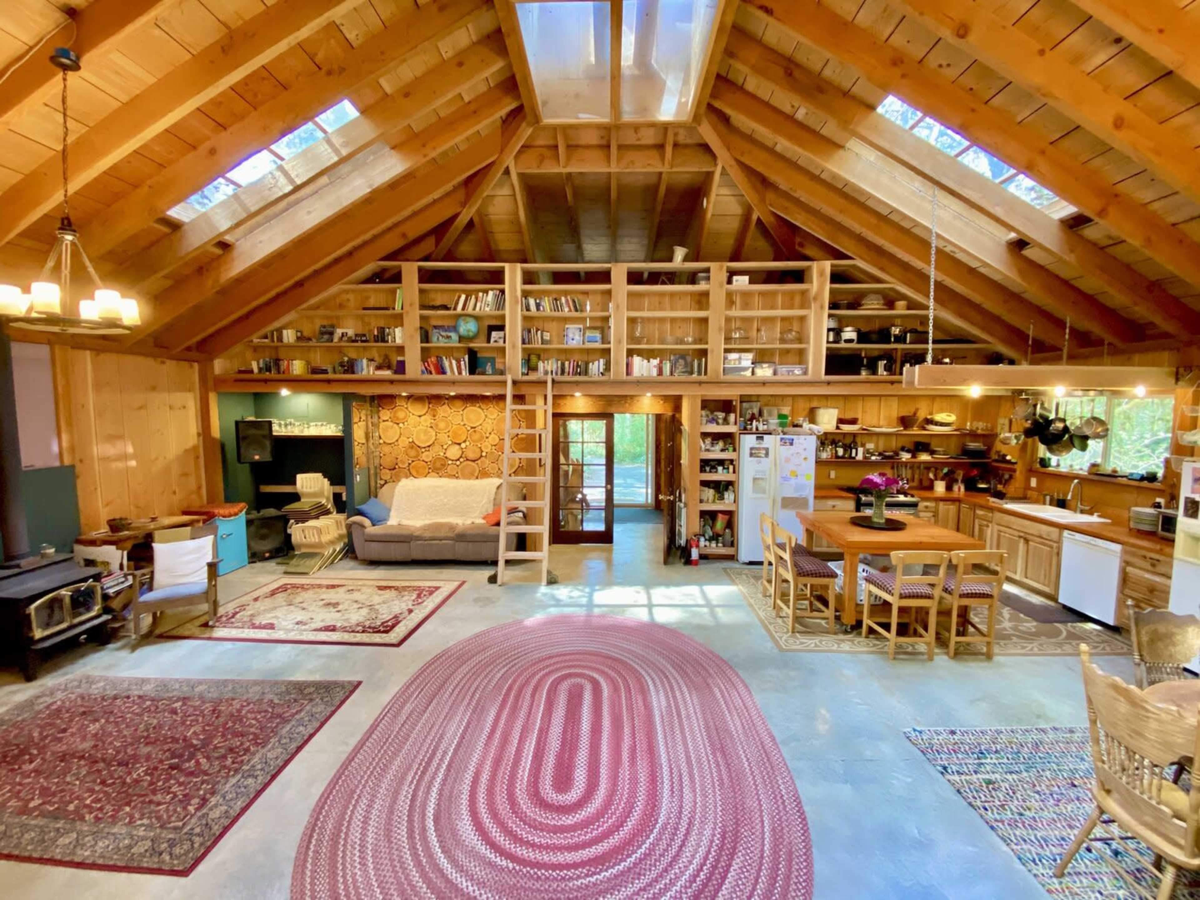 The image shows a spacious interior of a rustic cabin with wooden beams, bookshelves, a dining area, and a cozy seating area.