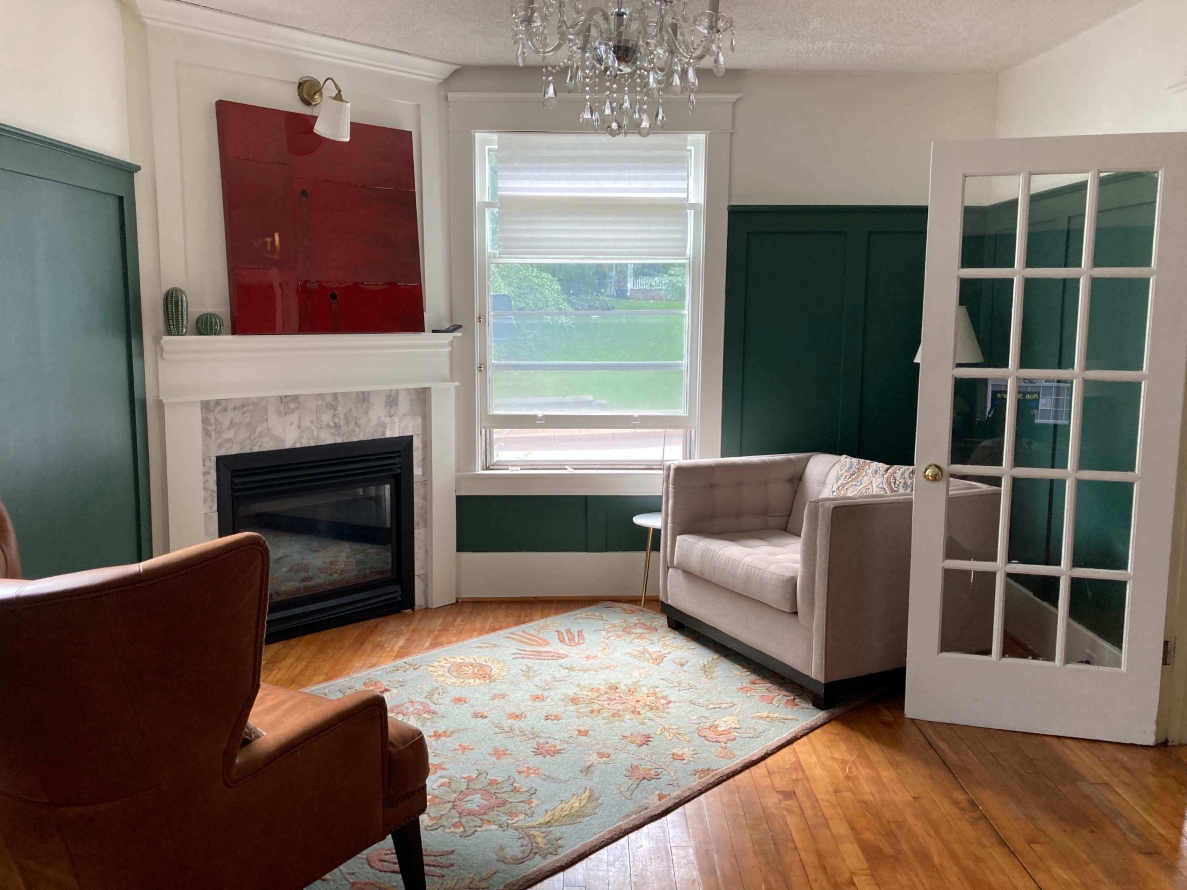 A cozy living room features a fireplace, green accent walls, a light-colored sofa, a brown armchair, and a decorative rug.