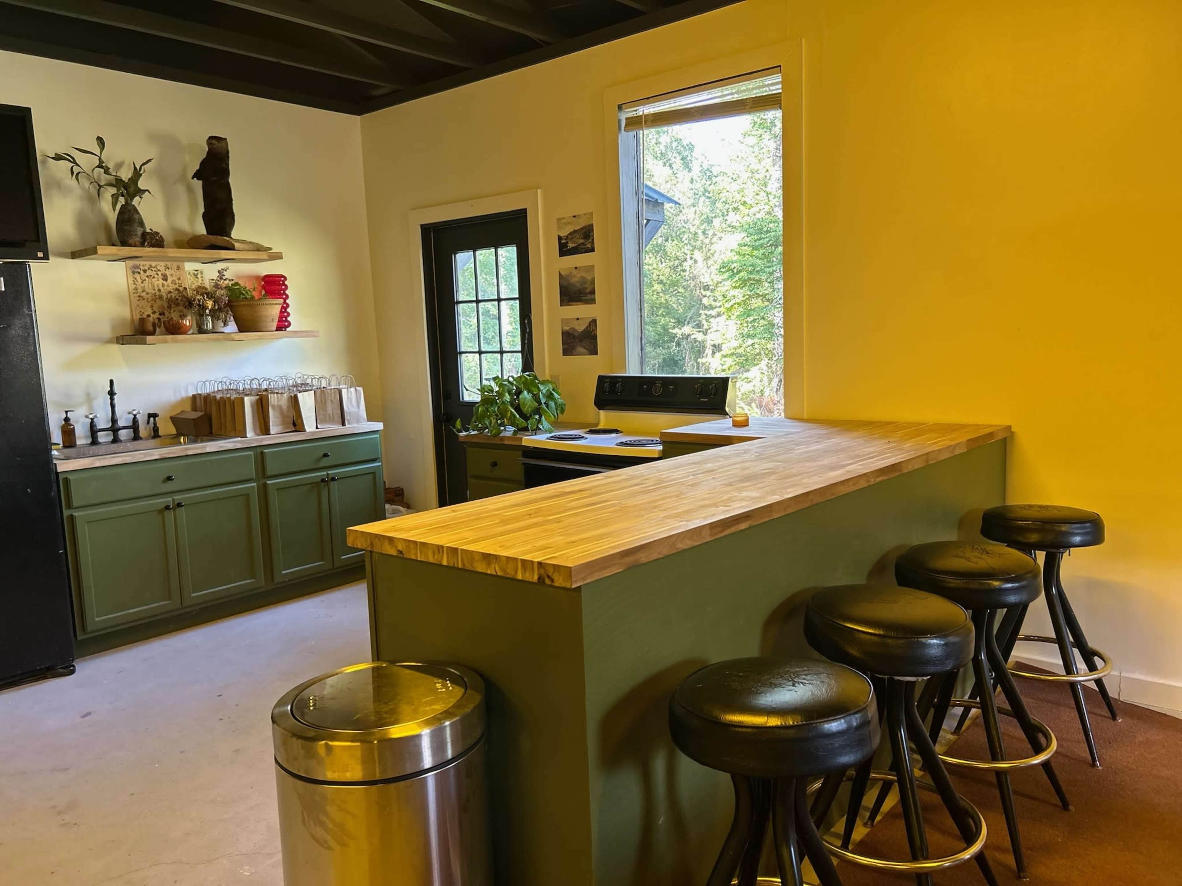 The image shows a kitchen area featuring a wooden counter with stools, green cabinetry, and a large window that allows natural light to enter.