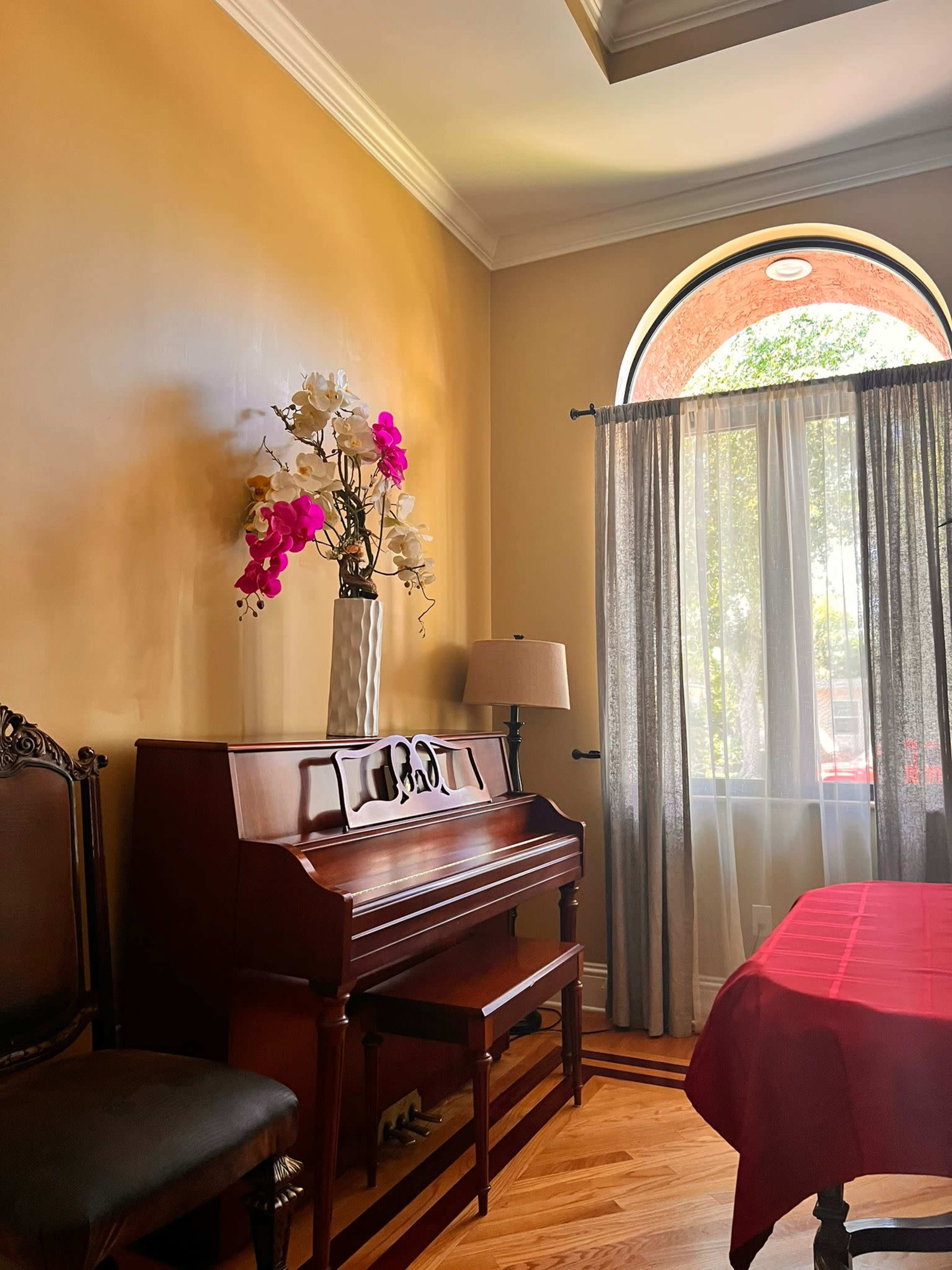 A wooden piano is positioned next to a window adorned with sheer curtains, with a vase of flowers on top and a lamp nearby.