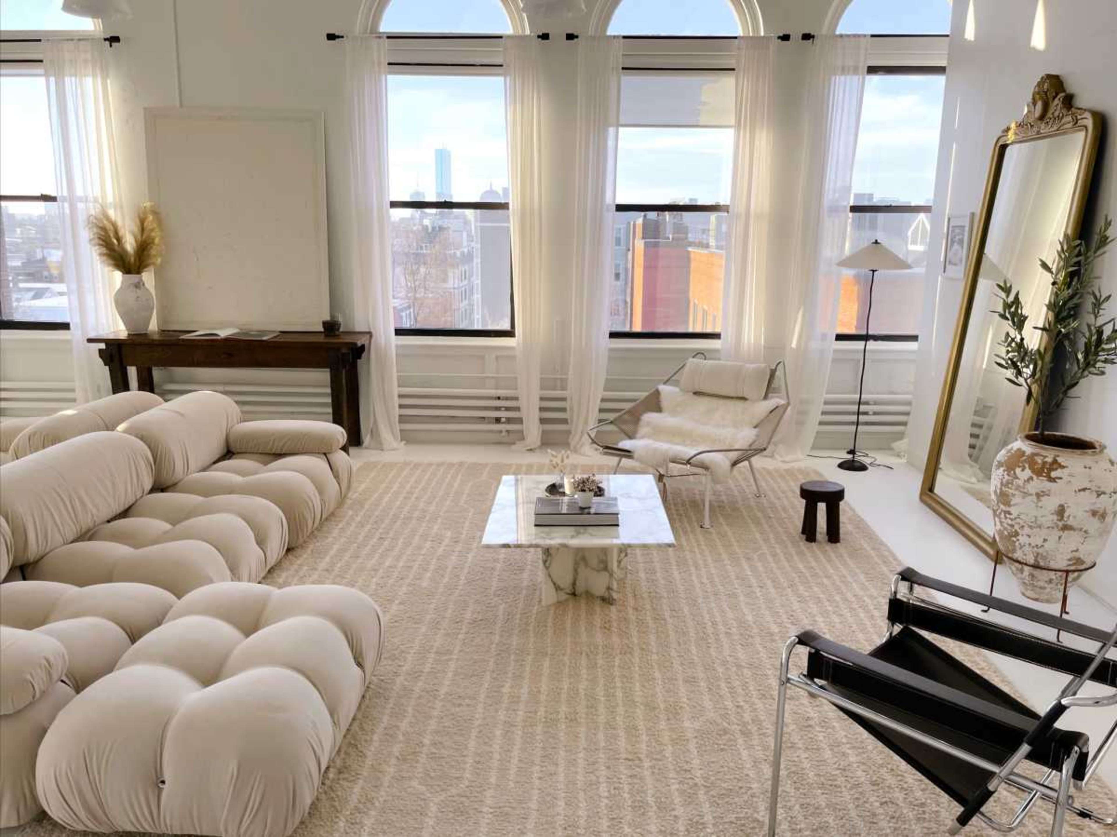 The room features a modern seating area with a beige tufted sofa, a marble coffee table, a minimalist chair, and large windows letting in natural light.