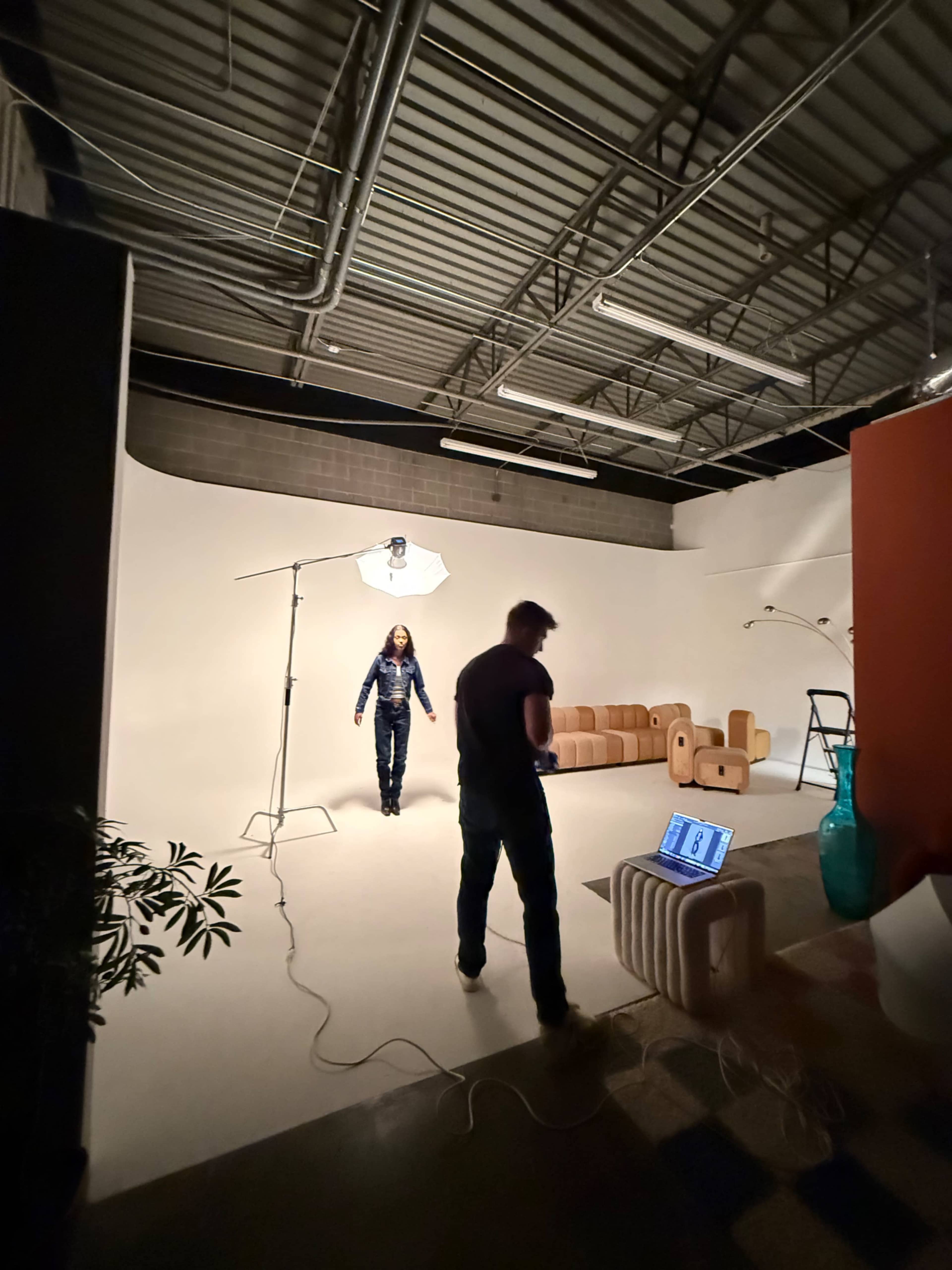 A model poses in a well-lit studio while a photographer adjusts equipment nearby.