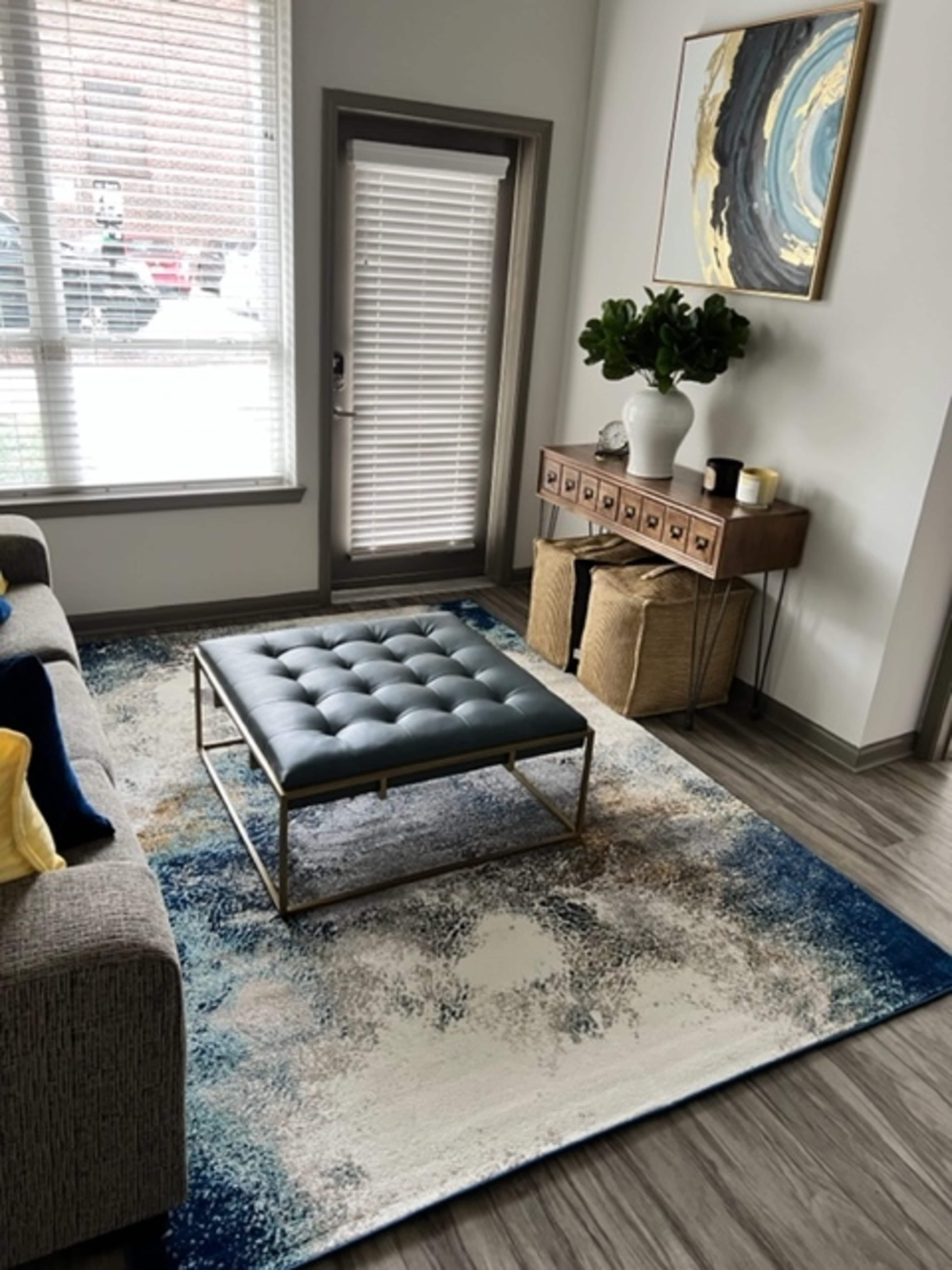 The image shows a modern living room with a gray sofa, a blue tufted ottoman on a decorative area rug, and a wooden side table with a plant and a decorative item.