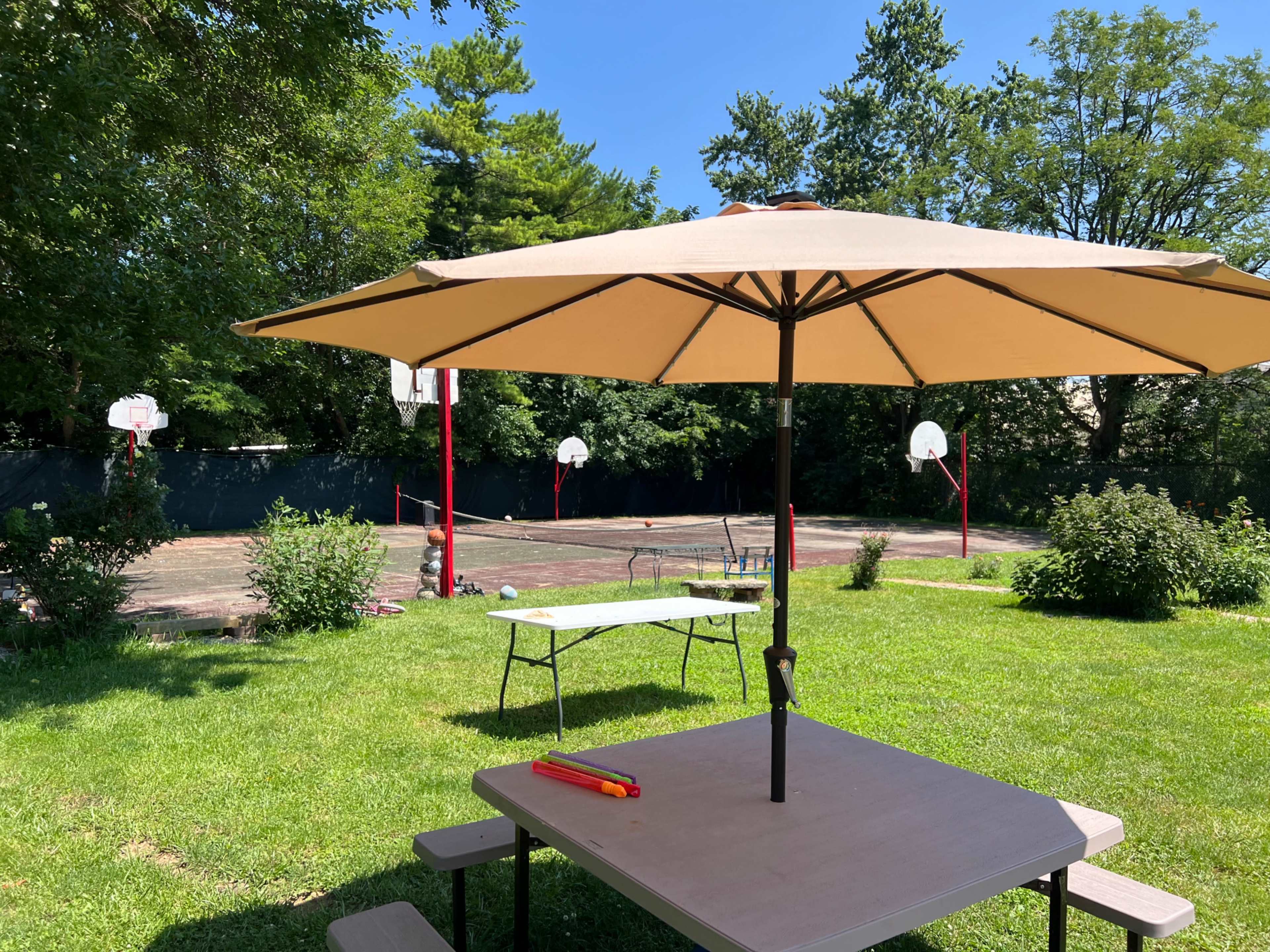 A shaded picnic table under a large umbrella overlooks a basketball court with several hoops and green trees in the background.