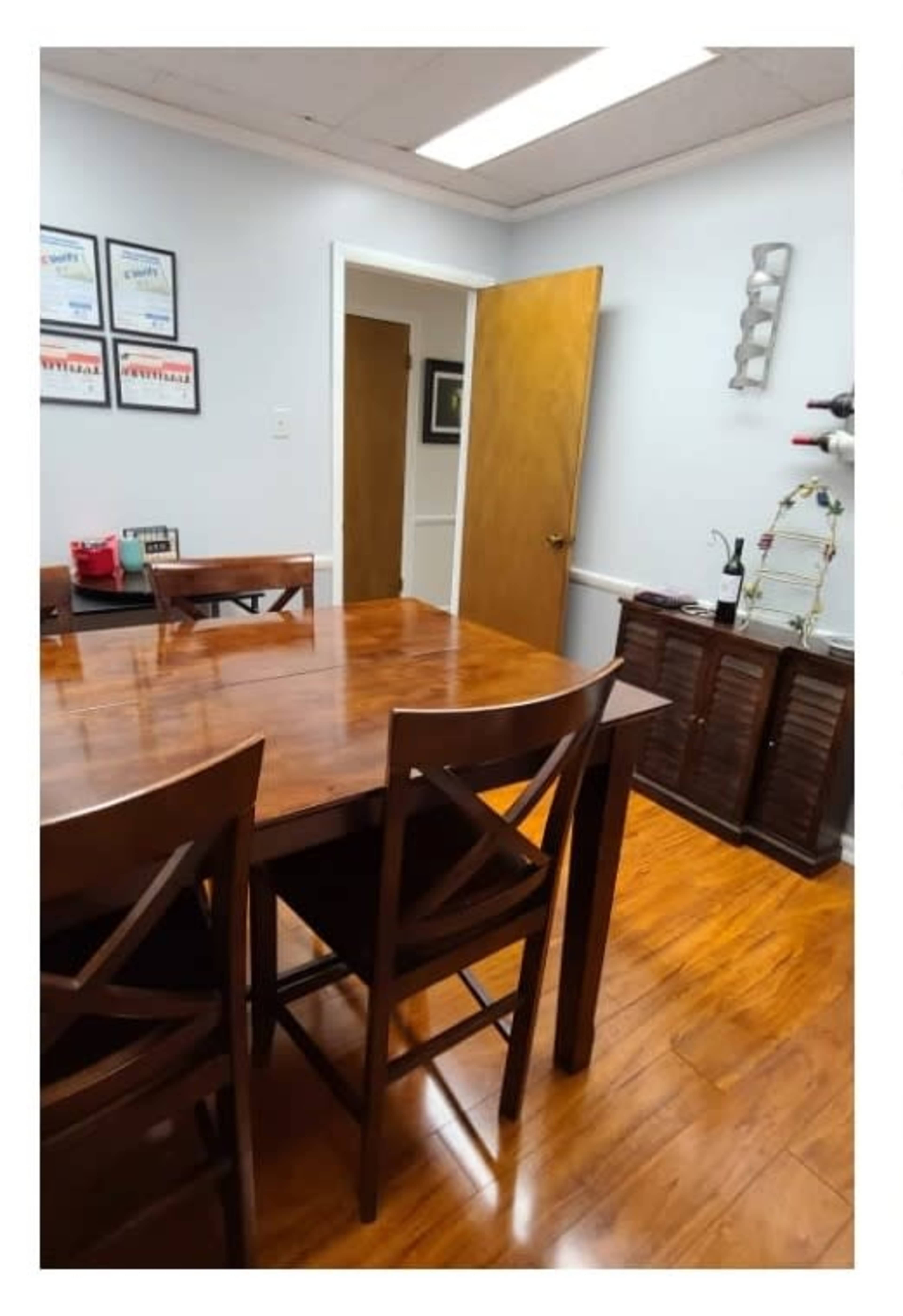 The image shows a dining area with a wooden table and chairs, framed certificates on the wall, and a partially open door leading to another room.