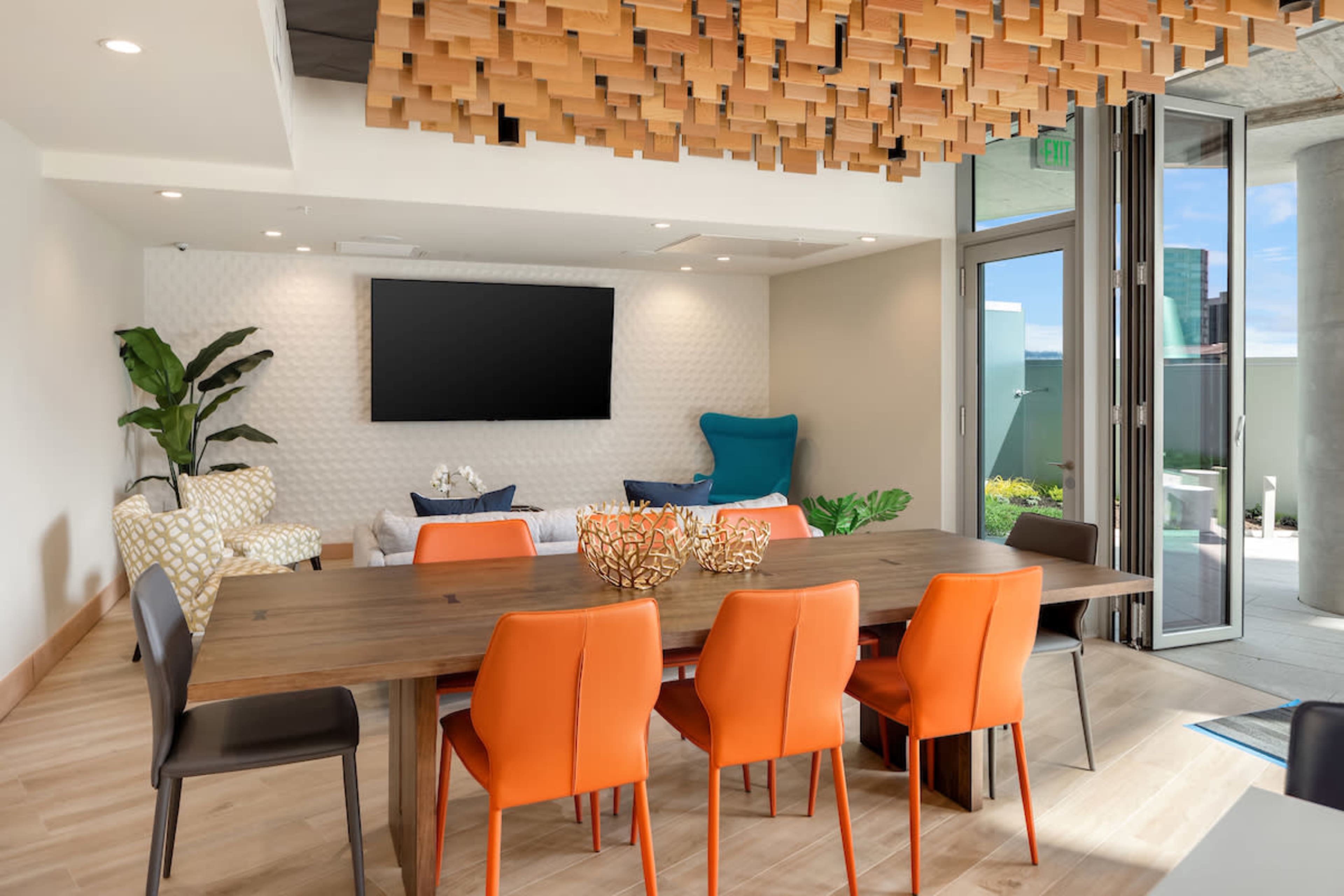 A modern meeting room features a large wooden table surrounded by orange and black chairs, with a television on the wall and large windows opening to an outdoor area.
