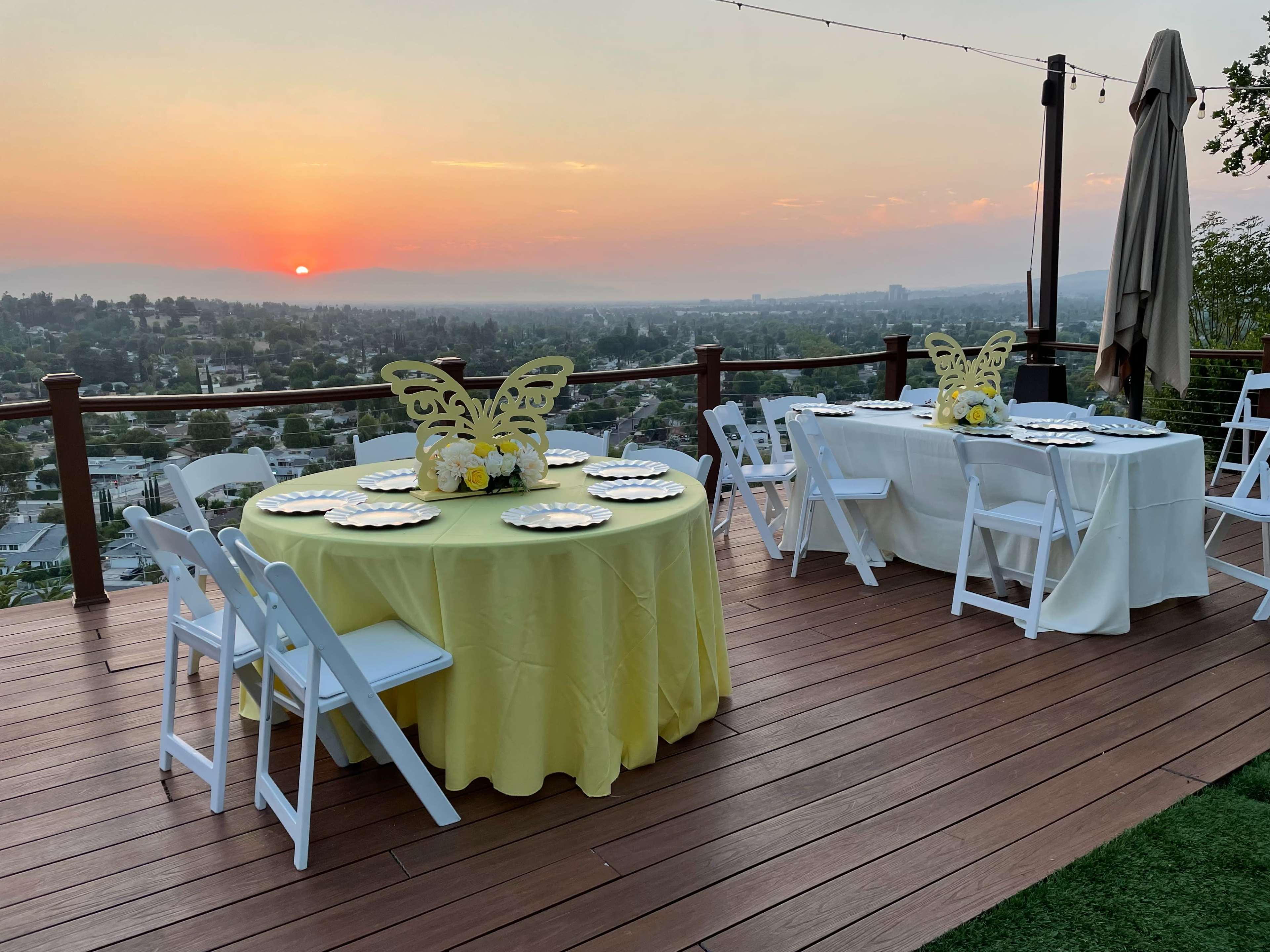 Two tables, one covered in yellow cloth and the other in white, are set for an outdoor event with plates arranged on each, against a backdrop of a sunset over a cityscape.