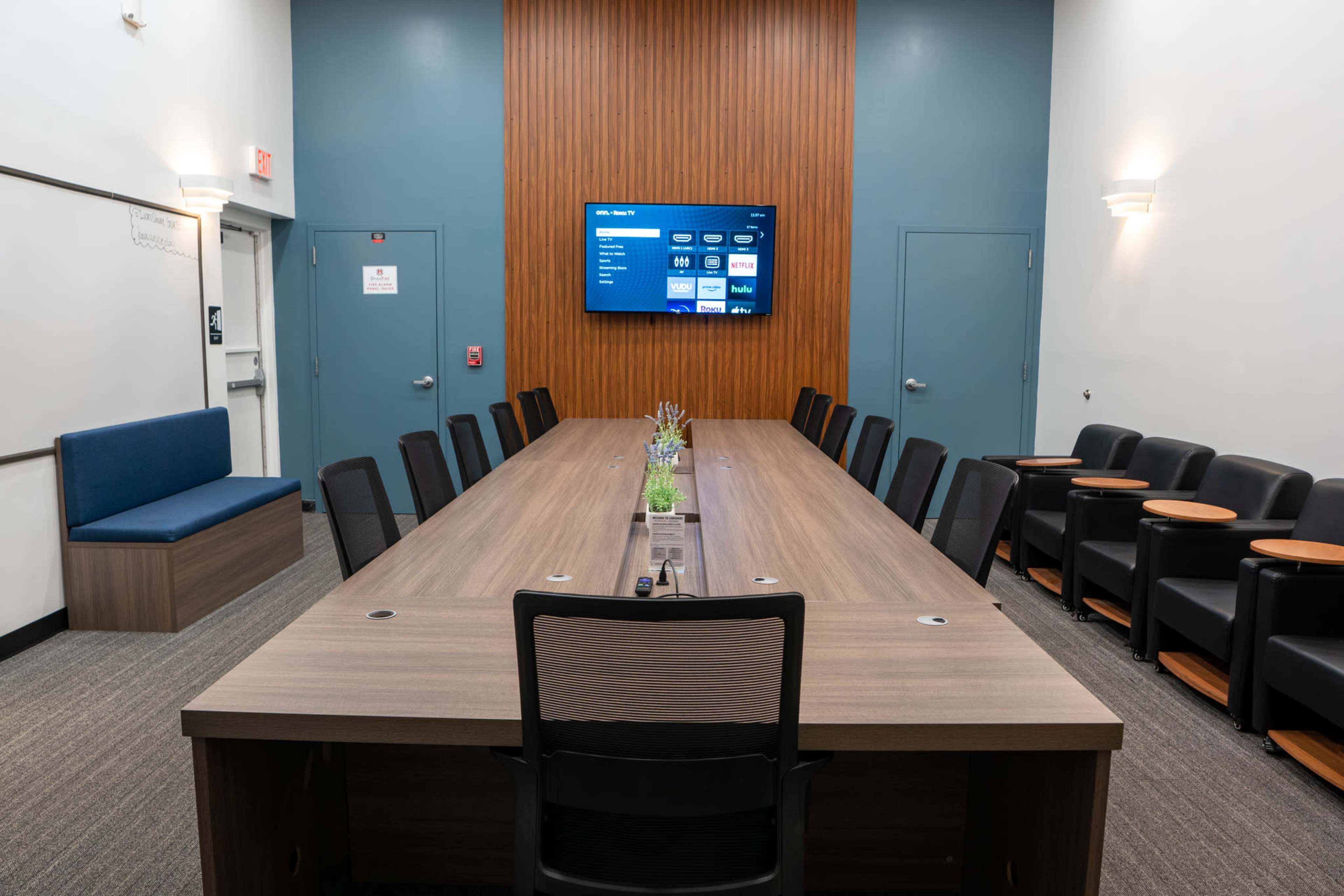 A long conference table with chairs surrounds a screen mounted on a wooden wall in a modern meeting room.