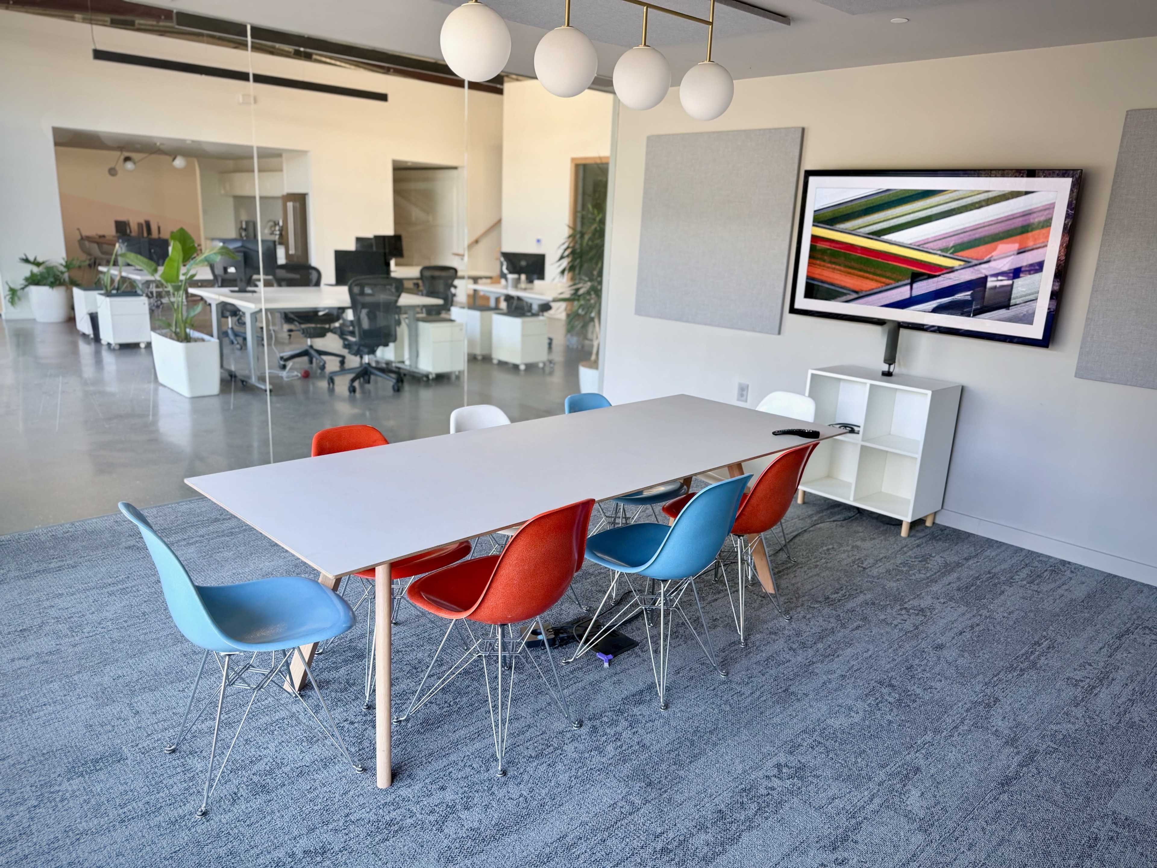 The image shows a modern conference room with a long rectangular table surrounded by colorful chairs, a television mounted on the wall, and a large window revealing an open office space in the background.