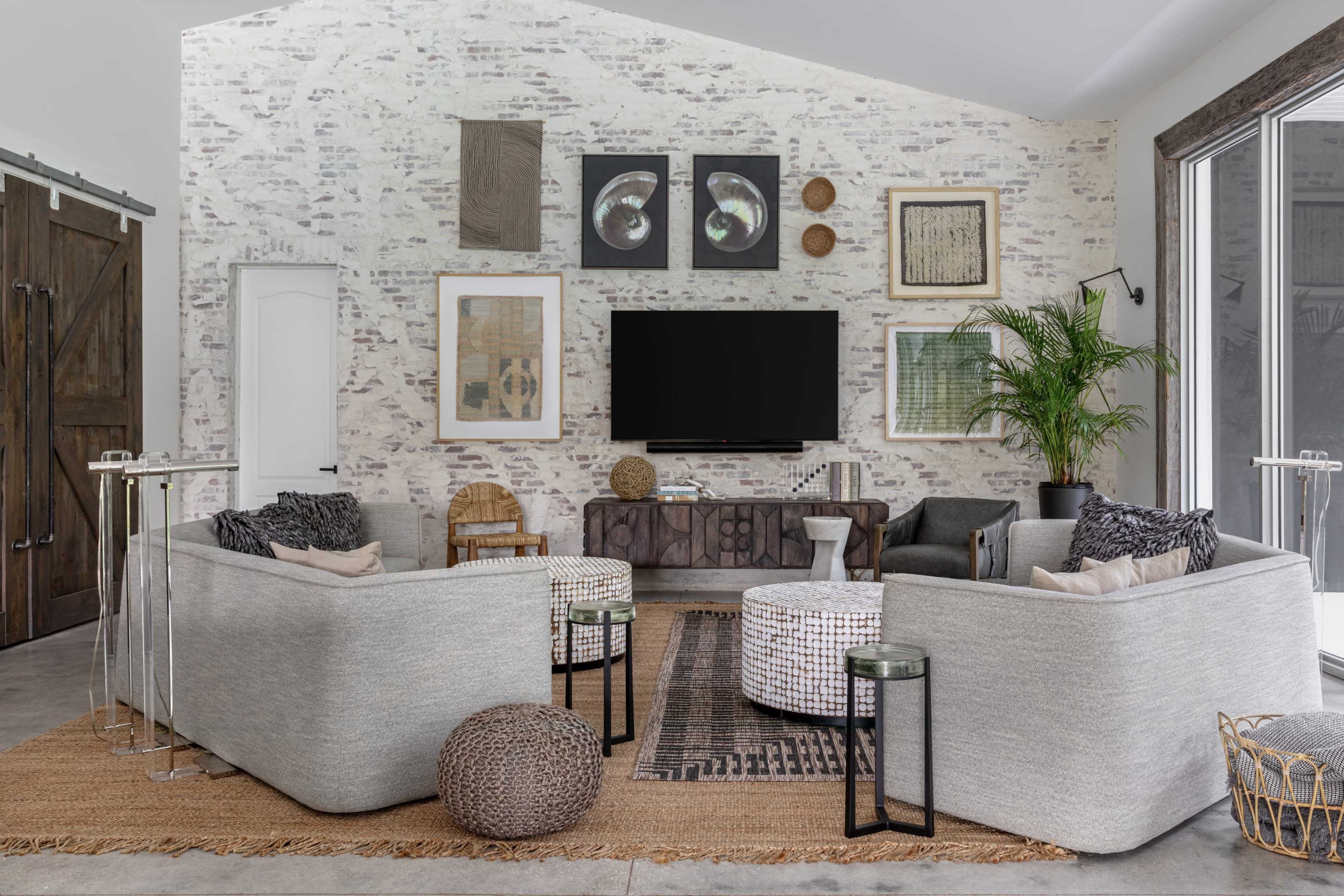 The image shows a modern living room featuring two gray sofas, a round coffee table, and decorative artworks on a bricked wall, with a large TV mounted above a wooden sideboard.