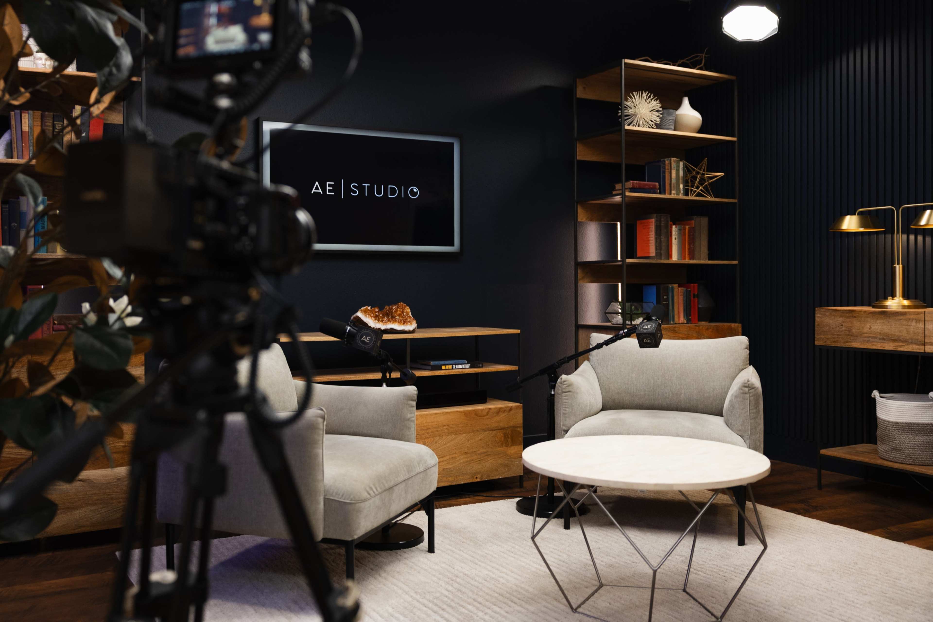The image shows a modern studio setup with two light grey chairs, a round coffee table, and a camera focused on a backdrop featuring a logo that reads "AE | STUDIO."