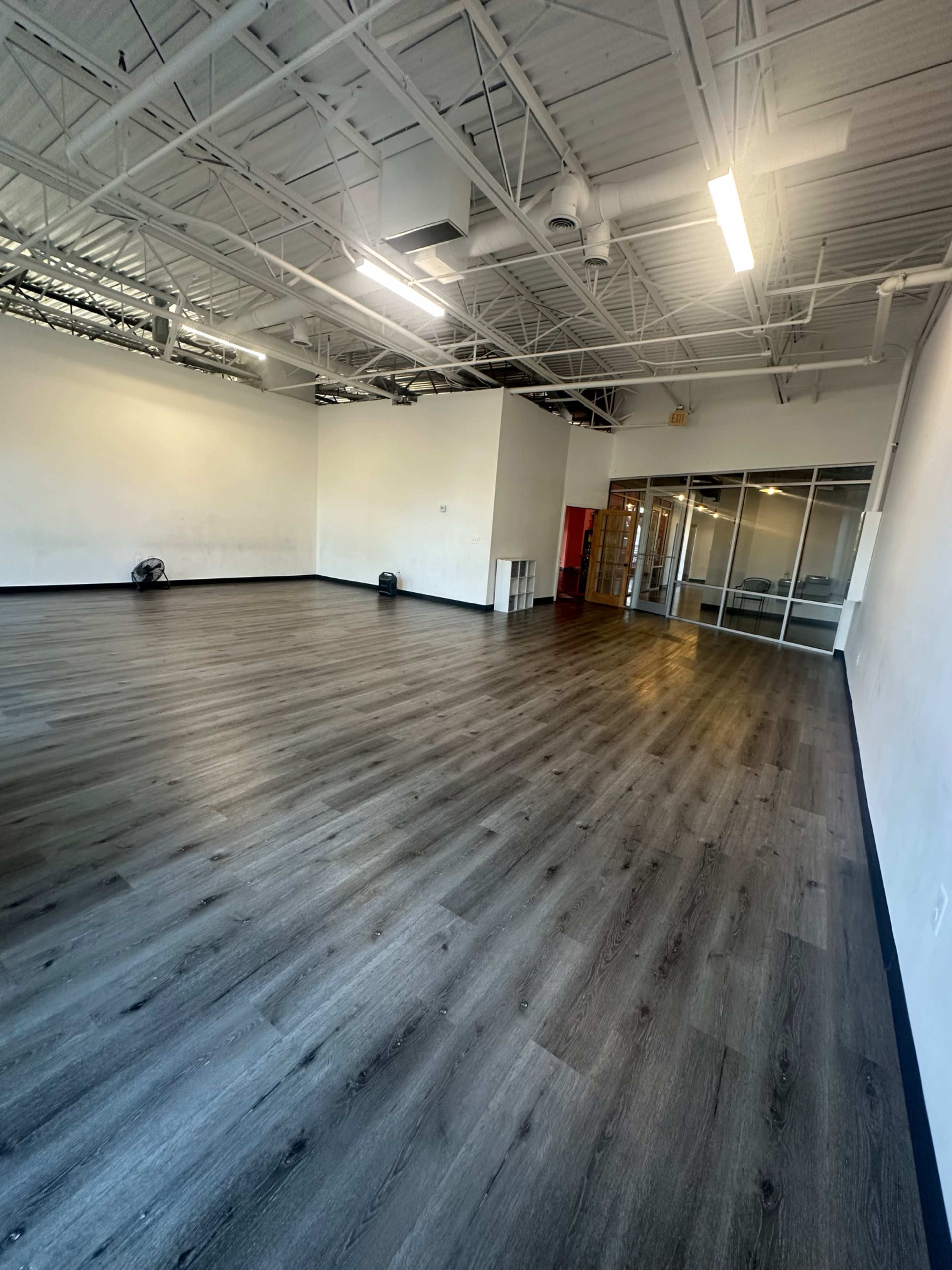 The image shows a spacious room with wooden flooring, white walls, and large windows, featuring a glass door at one end.