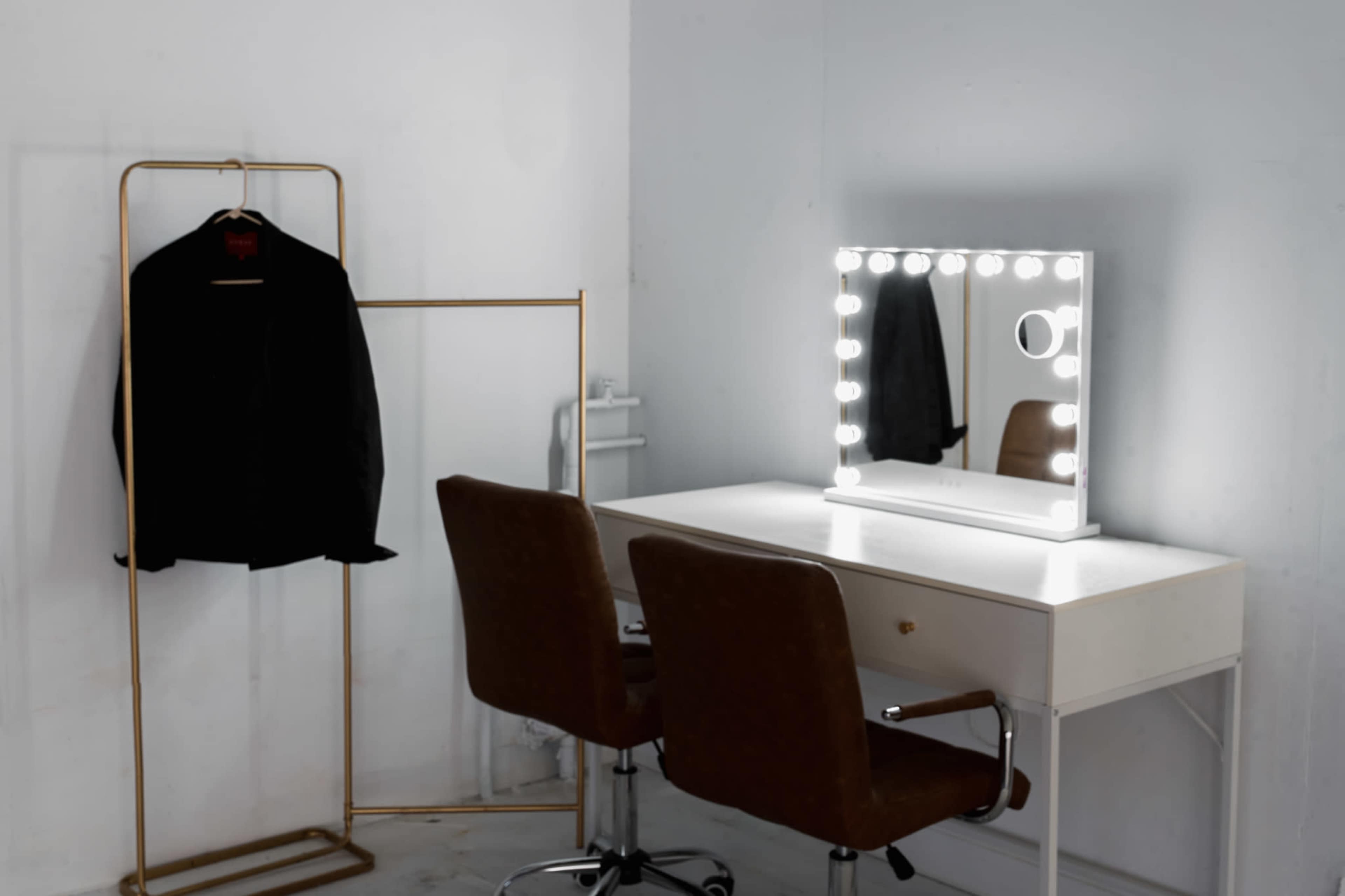 The image features a minimalist dressing area with a black garment hanging on a gold rack beside a white vanity table and illuminated mirror.