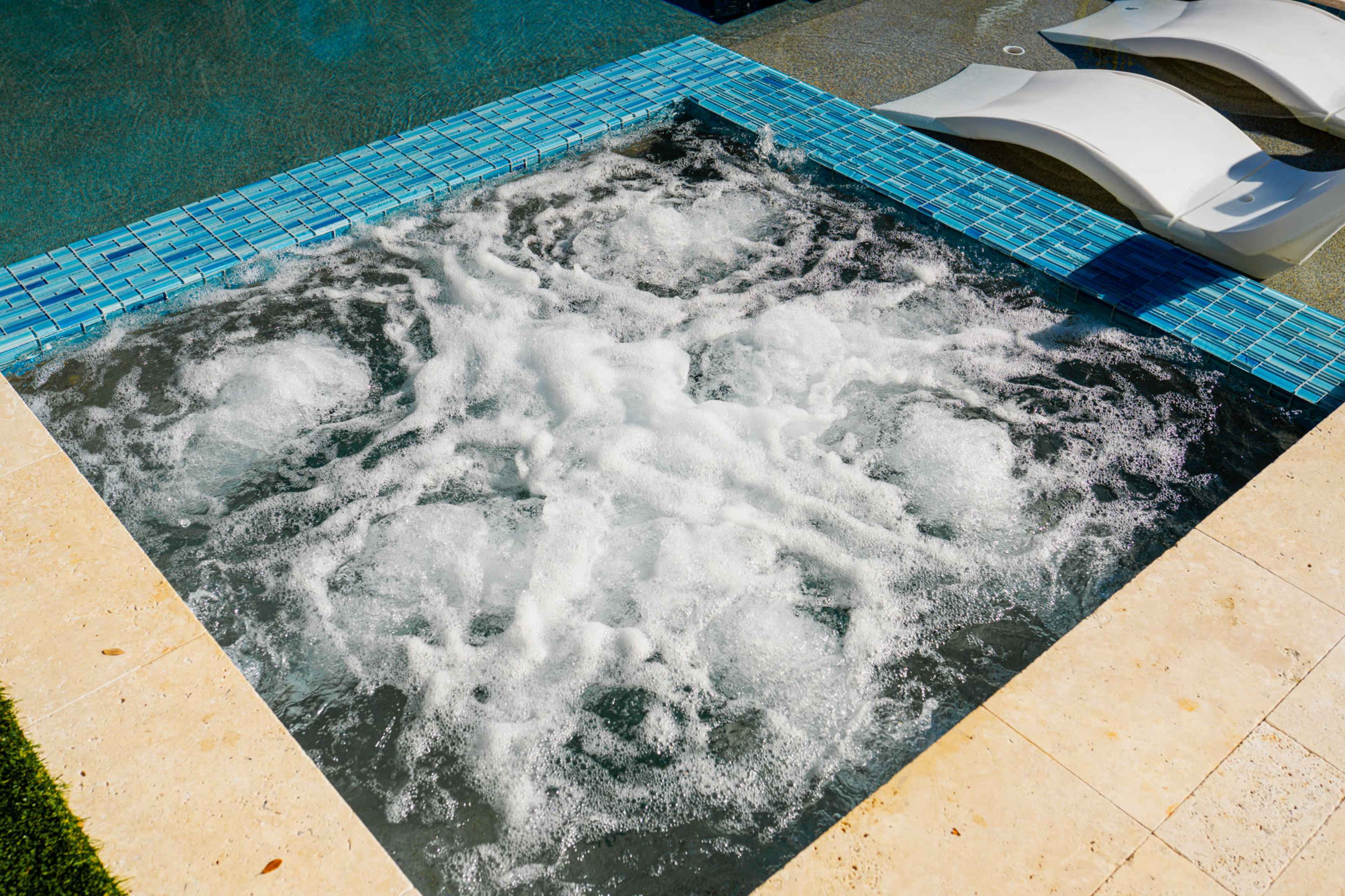 A bubbling hot tub is surrounded by a blue-tiled border and adjacent to lounge chairs by a swimming pool.