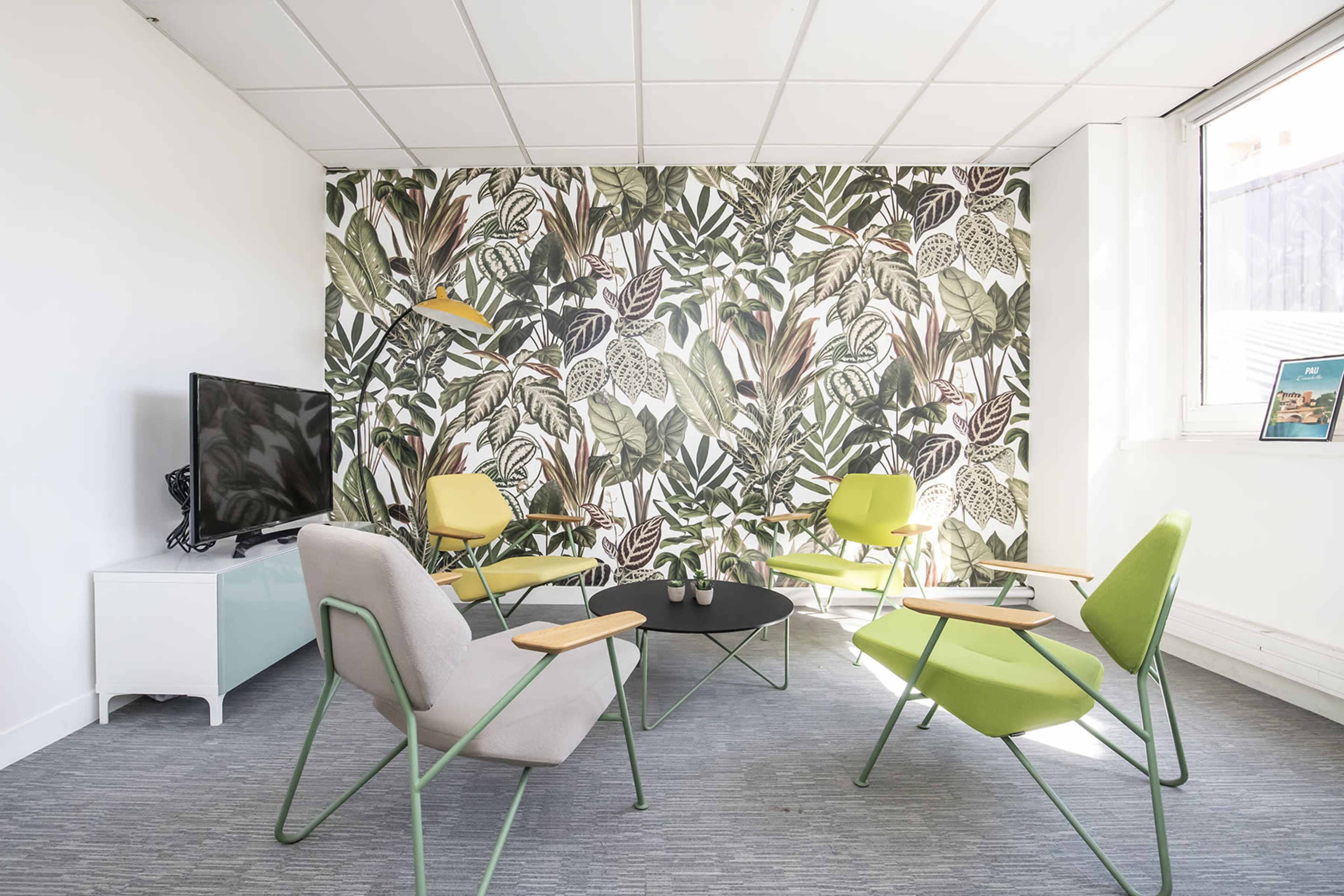 A modern room with a wall covered in a leafy plant design, featuring four green and gray chairs around a black coffee table and a television on a white stand.