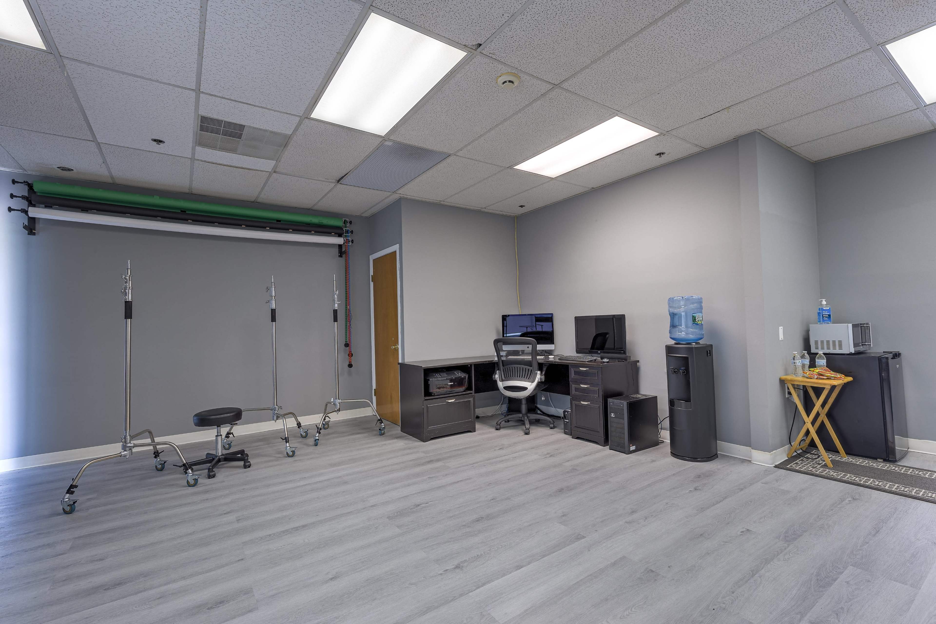 A spacious room with a desk and computer setup, a water cooler, and studio equipment stands against the walls.