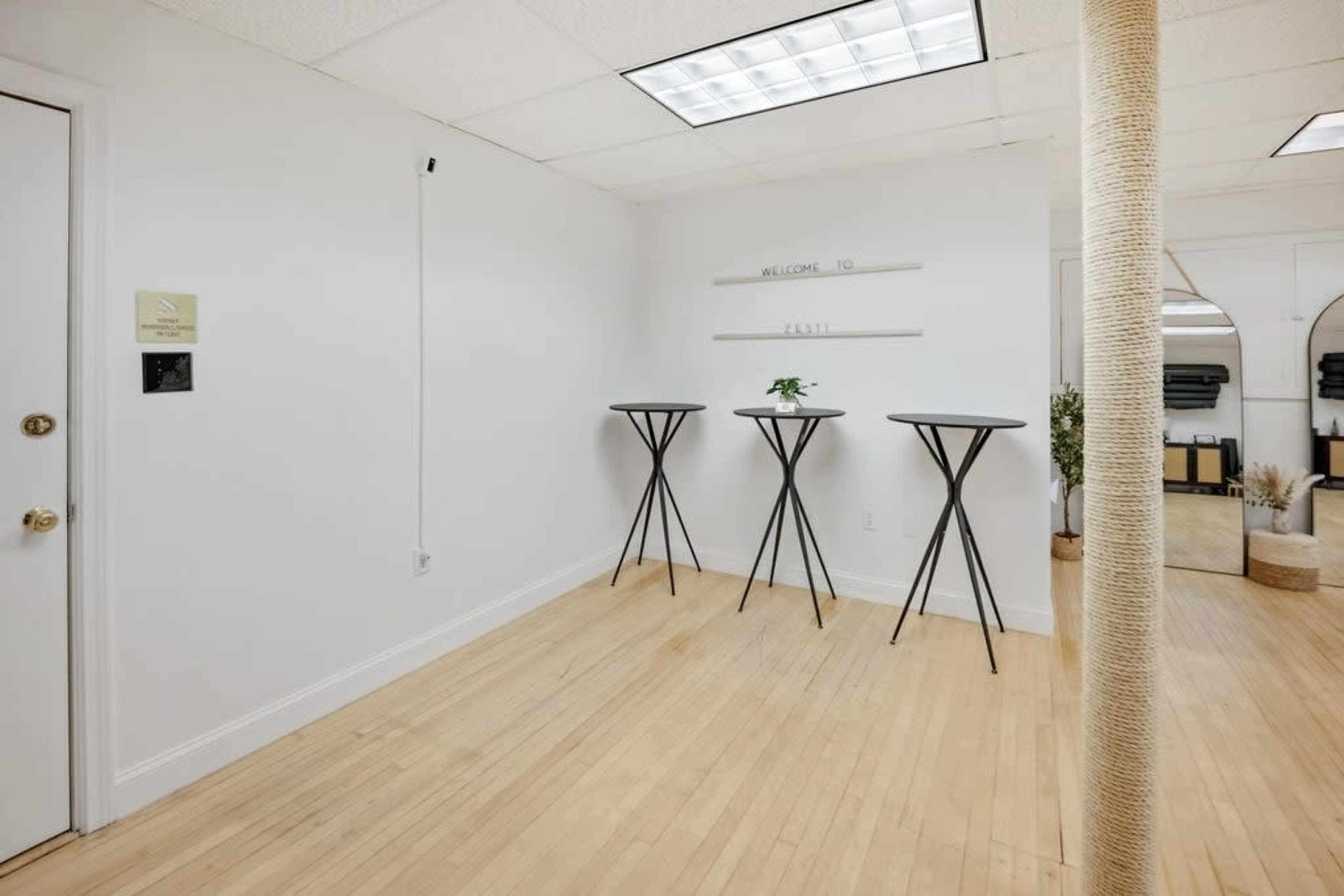 The room features three black high-top tables on a wooden floor, with a small plant on one table and mirrors along one wall.