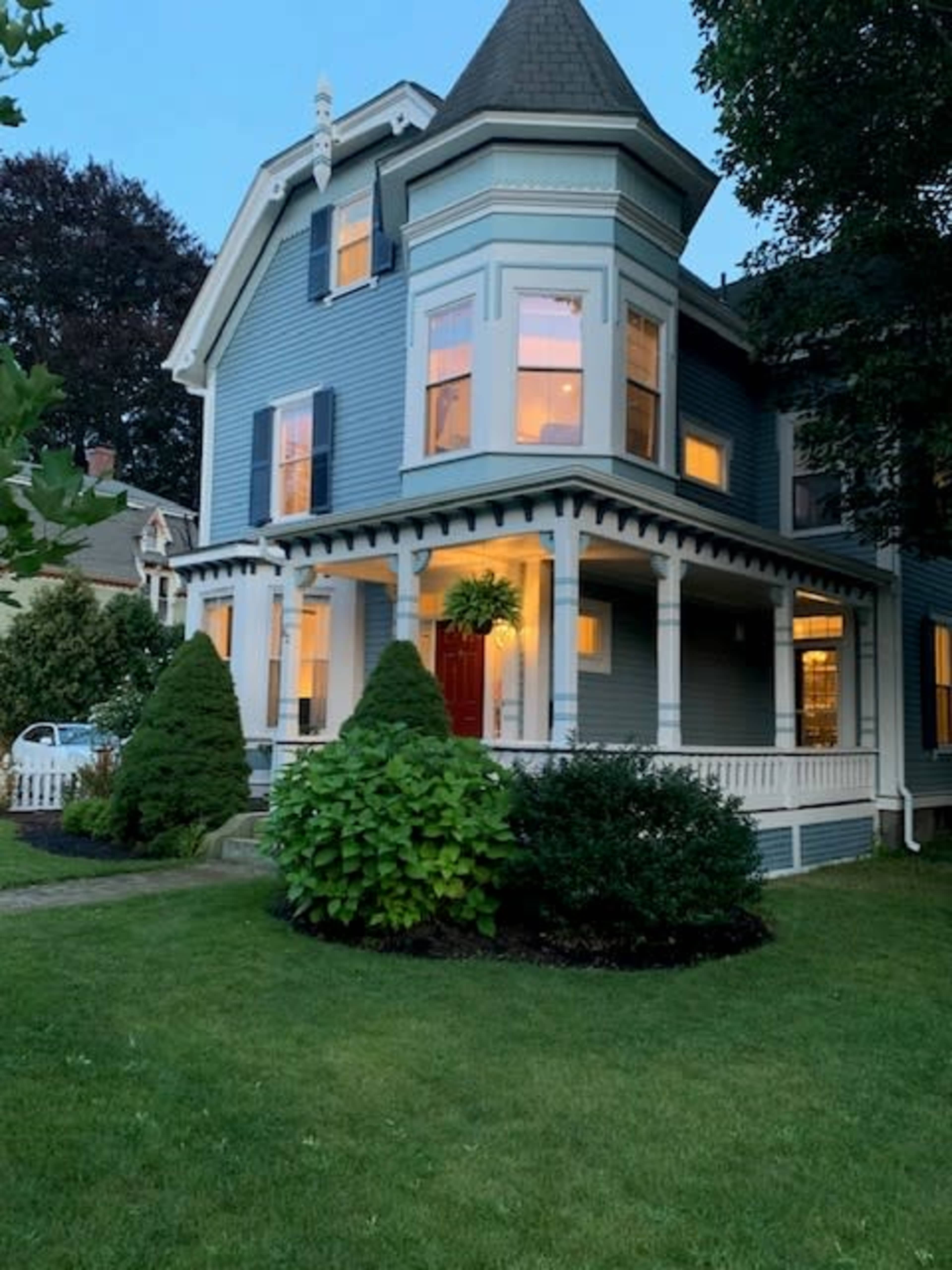 A blue Victorian house with a red front door, surrounded by lush greenery and illuminated windows at dusk.