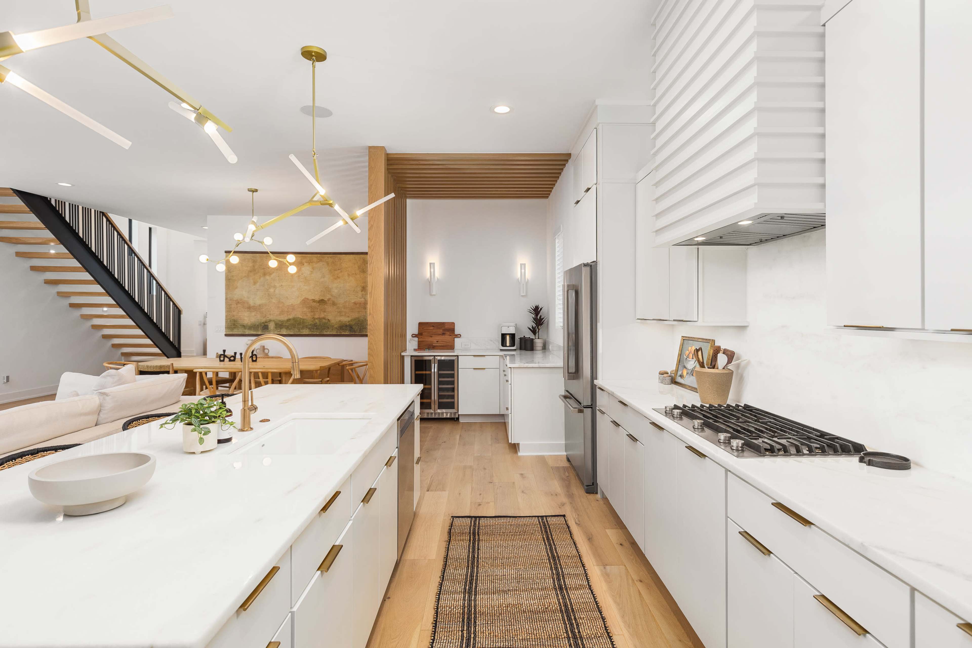 A modern kitchen features white cabinetry, marble countertops, a gas range, and a large island with a sink, with an open layout connecting to a living area and staircase.